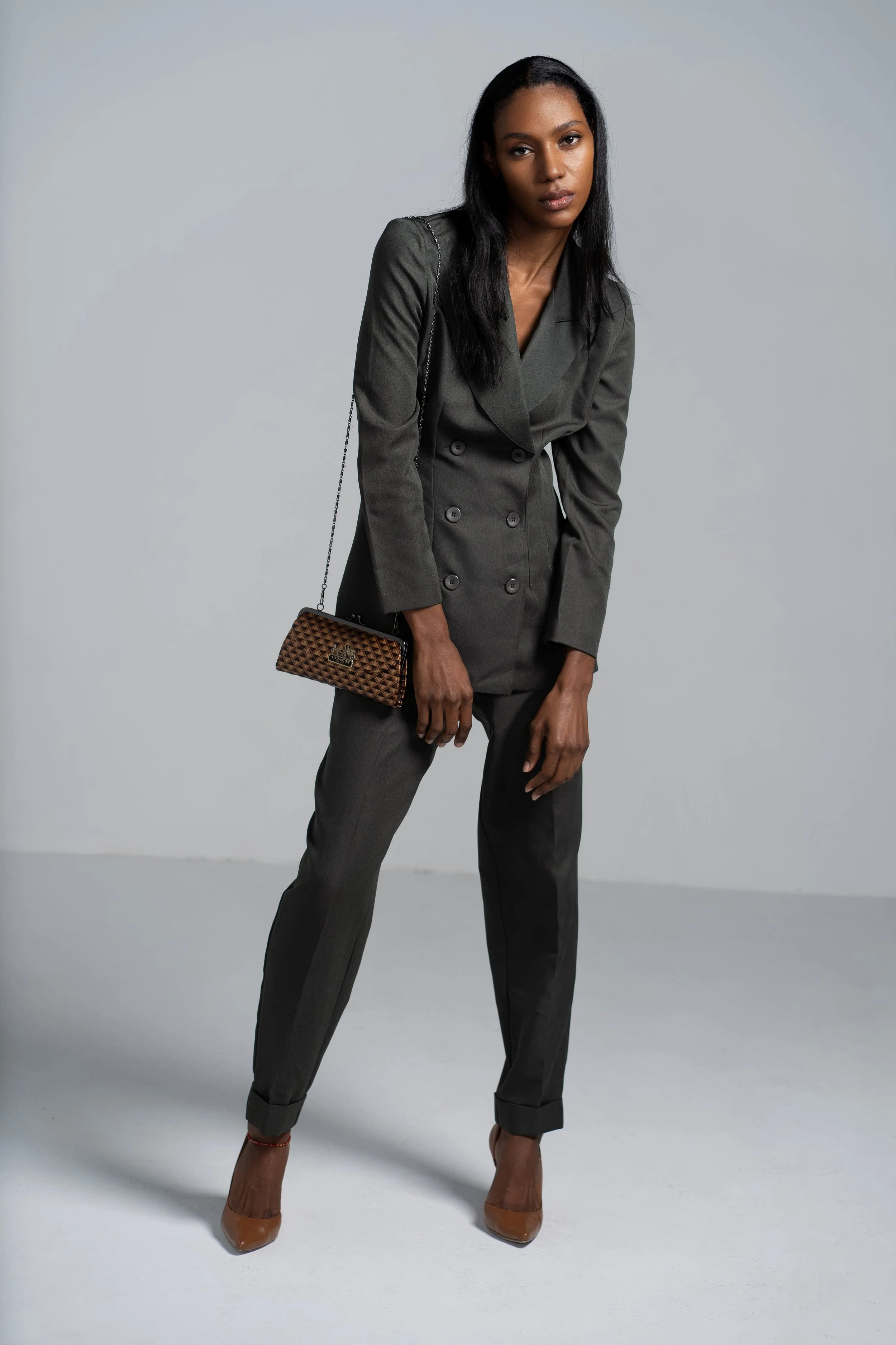 A woman with dark skin and long black hair wearing a tailored dark gray suit with trousers and a double-breasted blazer, brown high heels, and carrying a brown patterned clutch purse, standing against a plain gray background.