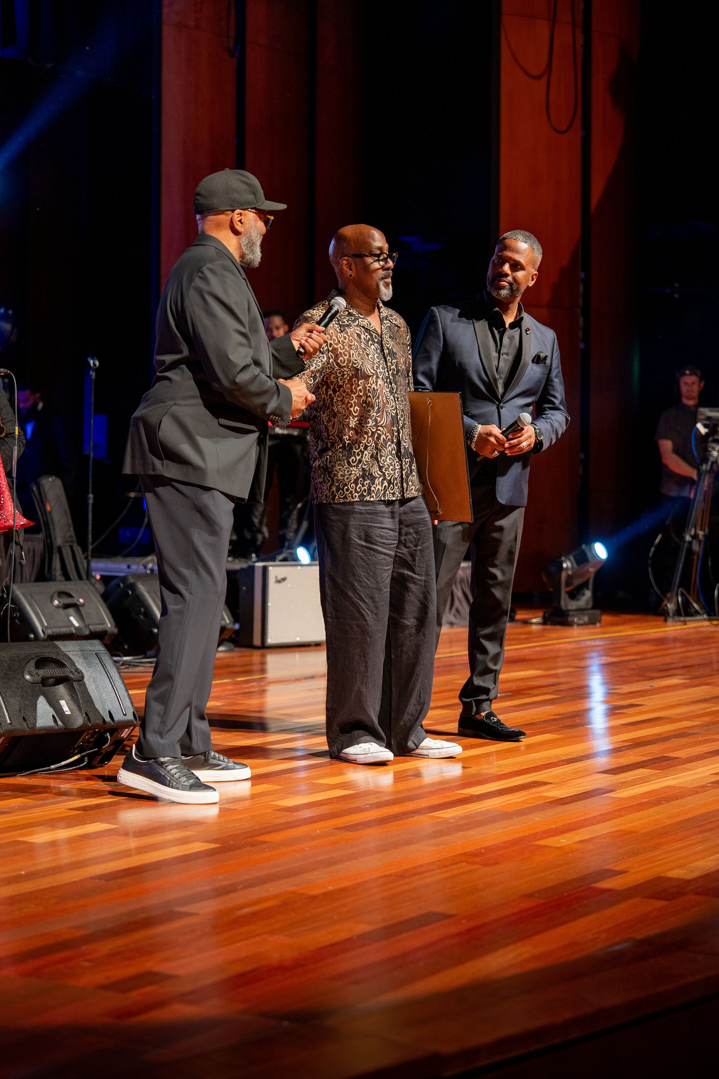 Three men standing on a stage in conversation, with music equipment and wooden paneling in the background.