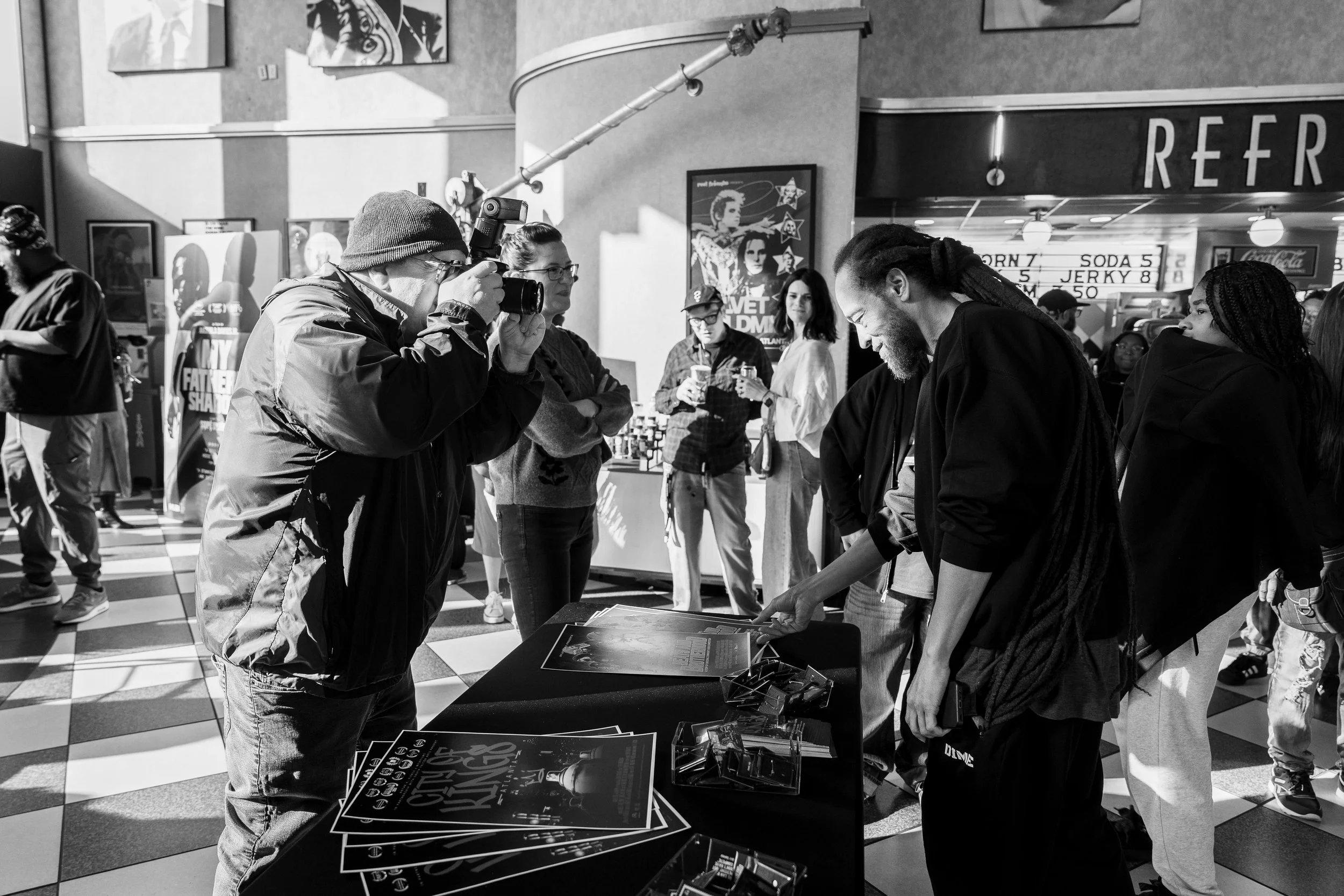 People at a convention or event, watching a man take a photo of another man who is signing posters at a booth.