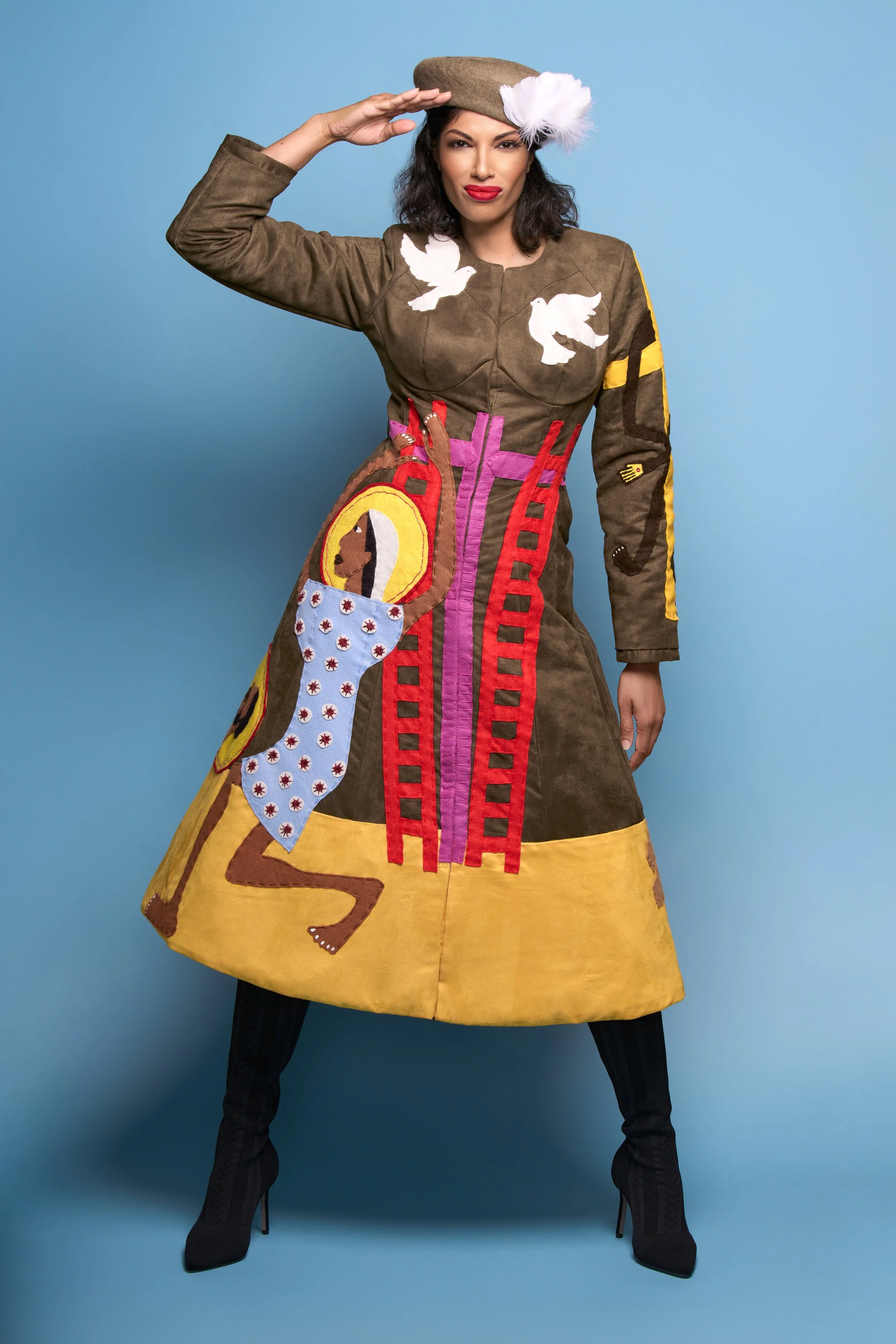 Woman wearing a colorful patchwork dress with white doves, a lion face, and a pocket with red and white patterns, accessorized with a beige hat with a white feather, posing against a blue background.