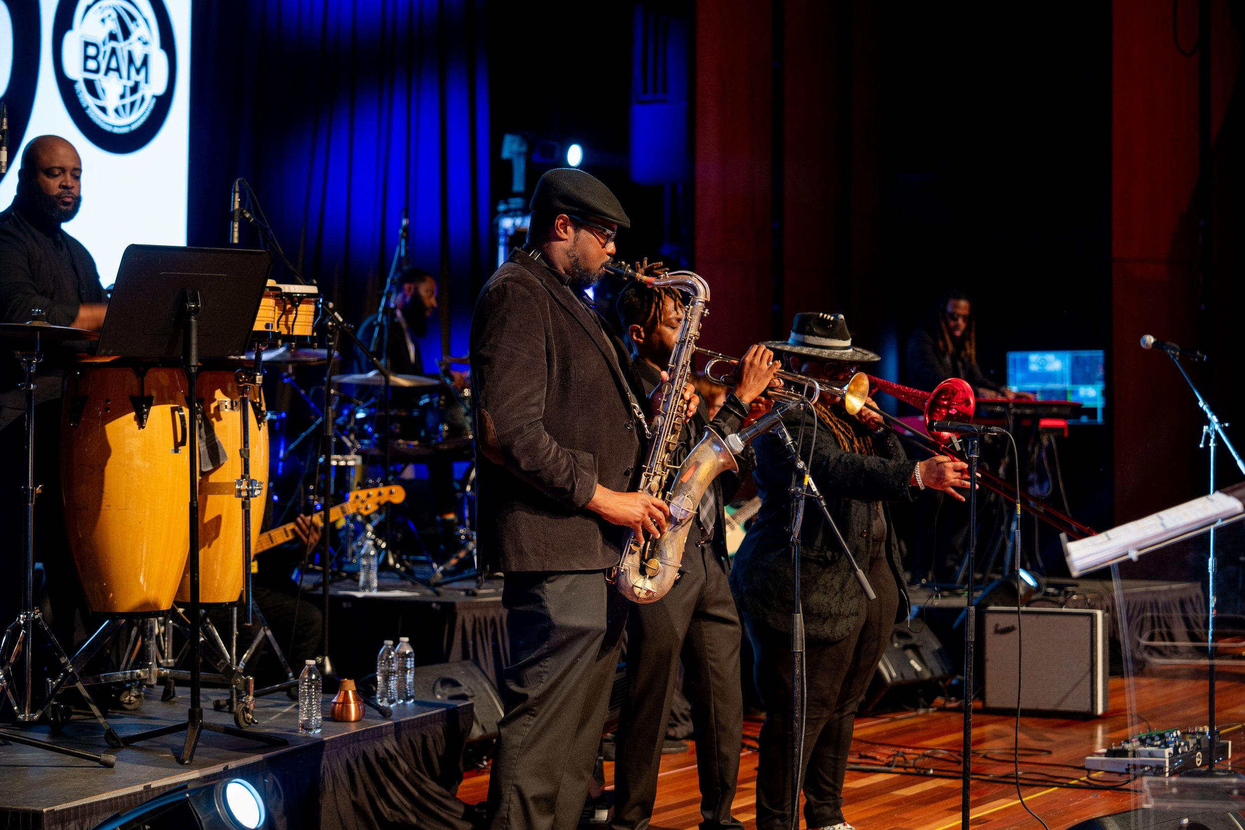 A group of musicians performing on a stage, including a man playing the saxophone, with others playing various instruments. There is a logo projected on a screen behind them.