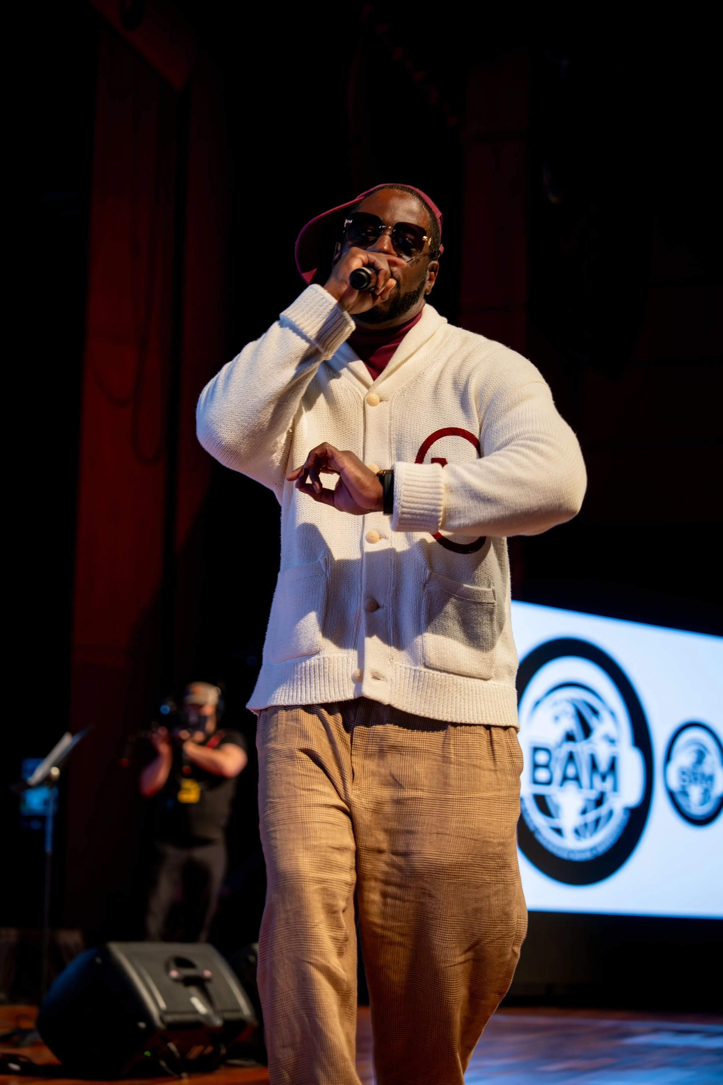 Man wearing sunglasses, red cap, and cream cardigan singing into microphones on stage with a logo in the background.