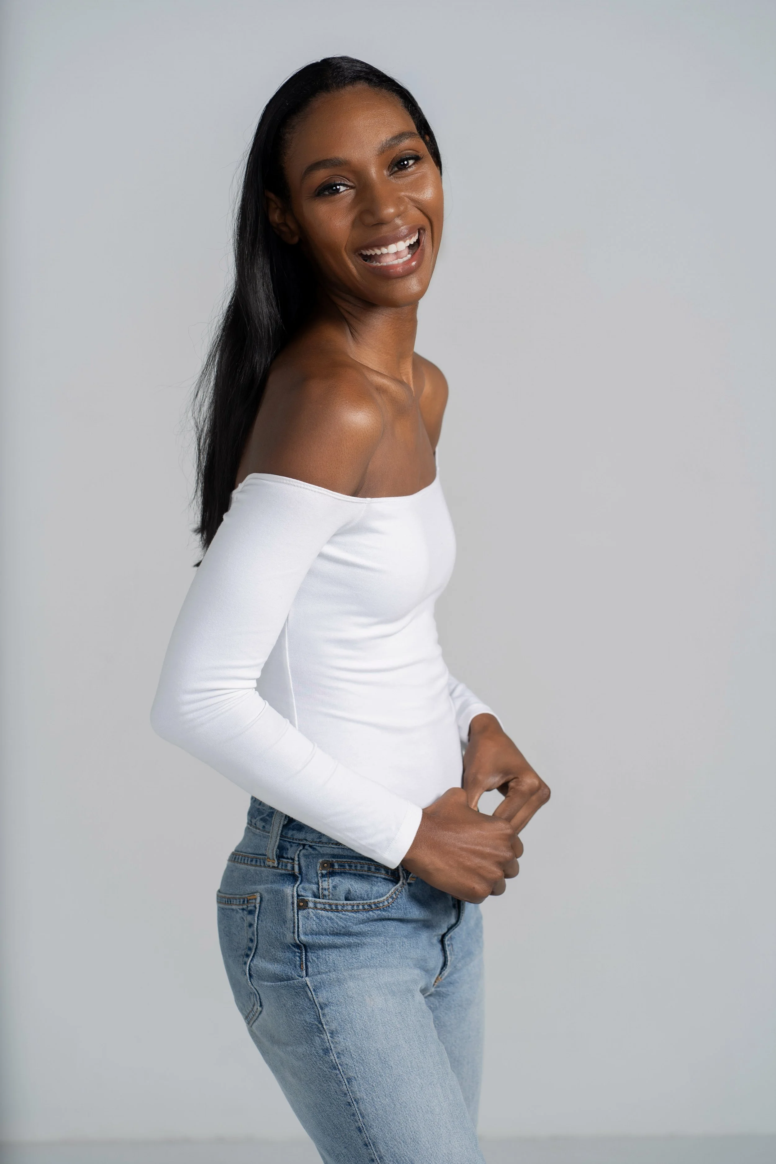 A woman with dark skin and long black hair, wearing a white off-the-shoulder top and blue jeans, smiling and posing against a plain light gray background.