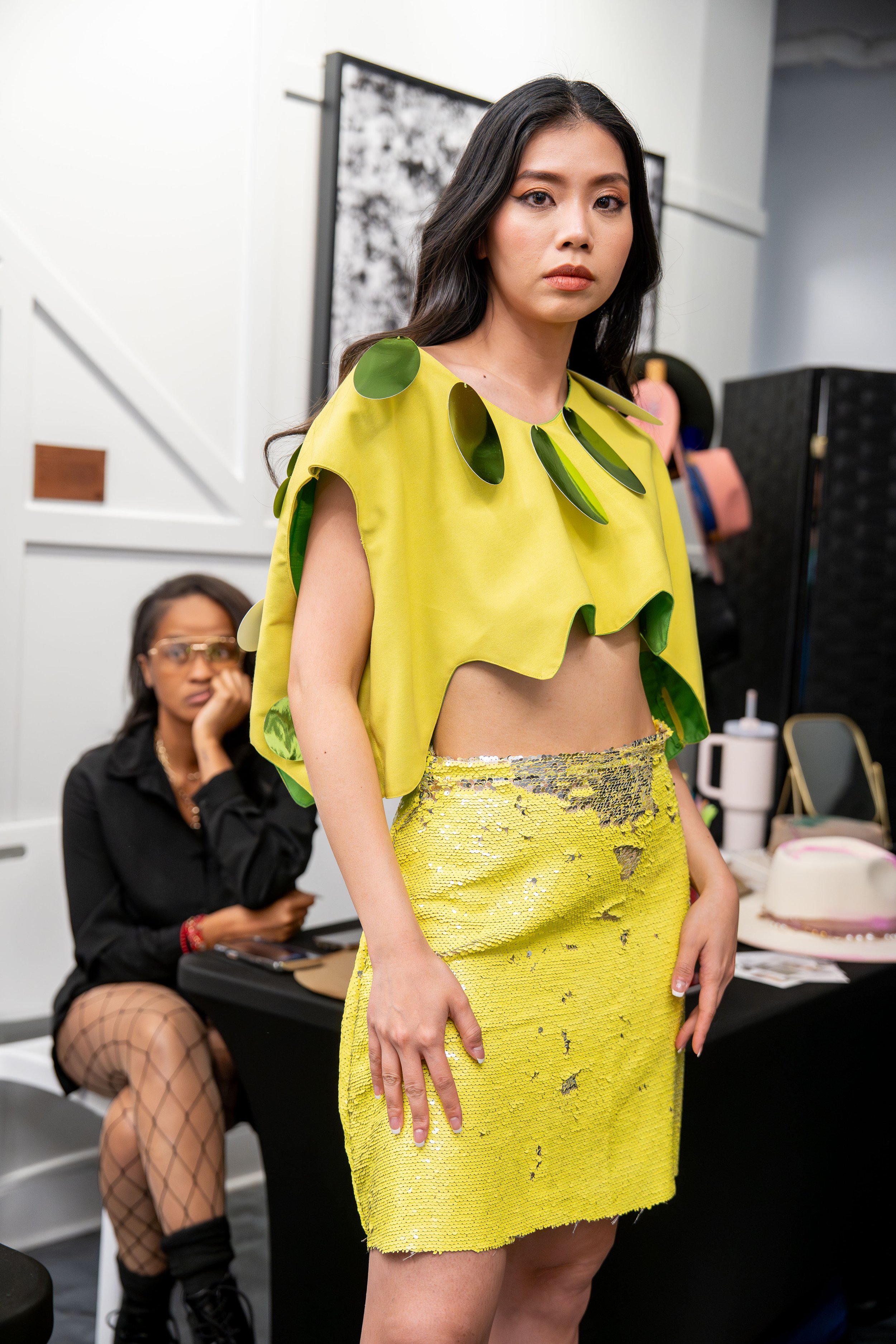 A woman in a yellow dress with green leaf embellishments standing in a room, with two women seated behind her; one with dark hair and glasses and another wearing fishnet stockings, sitting at a table.