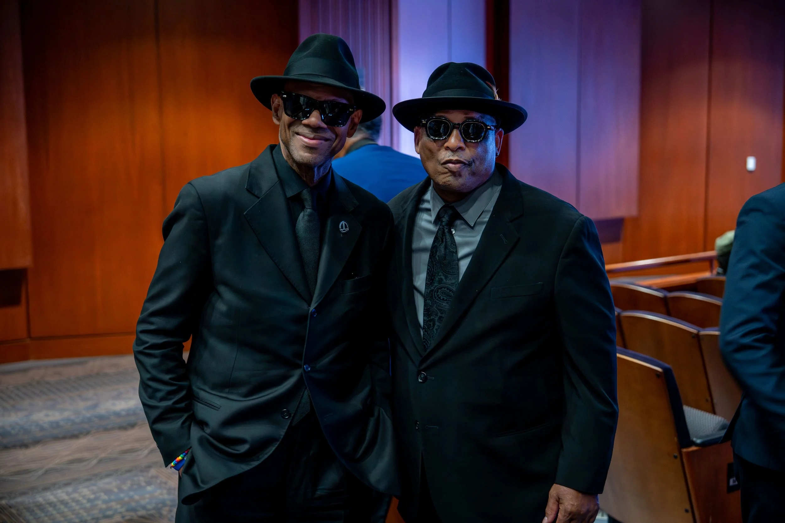Two men dressed in black suits, black hats, and sunglasses smiling at the camera. They are standing inside a room with wooden paneling and blue lighting.