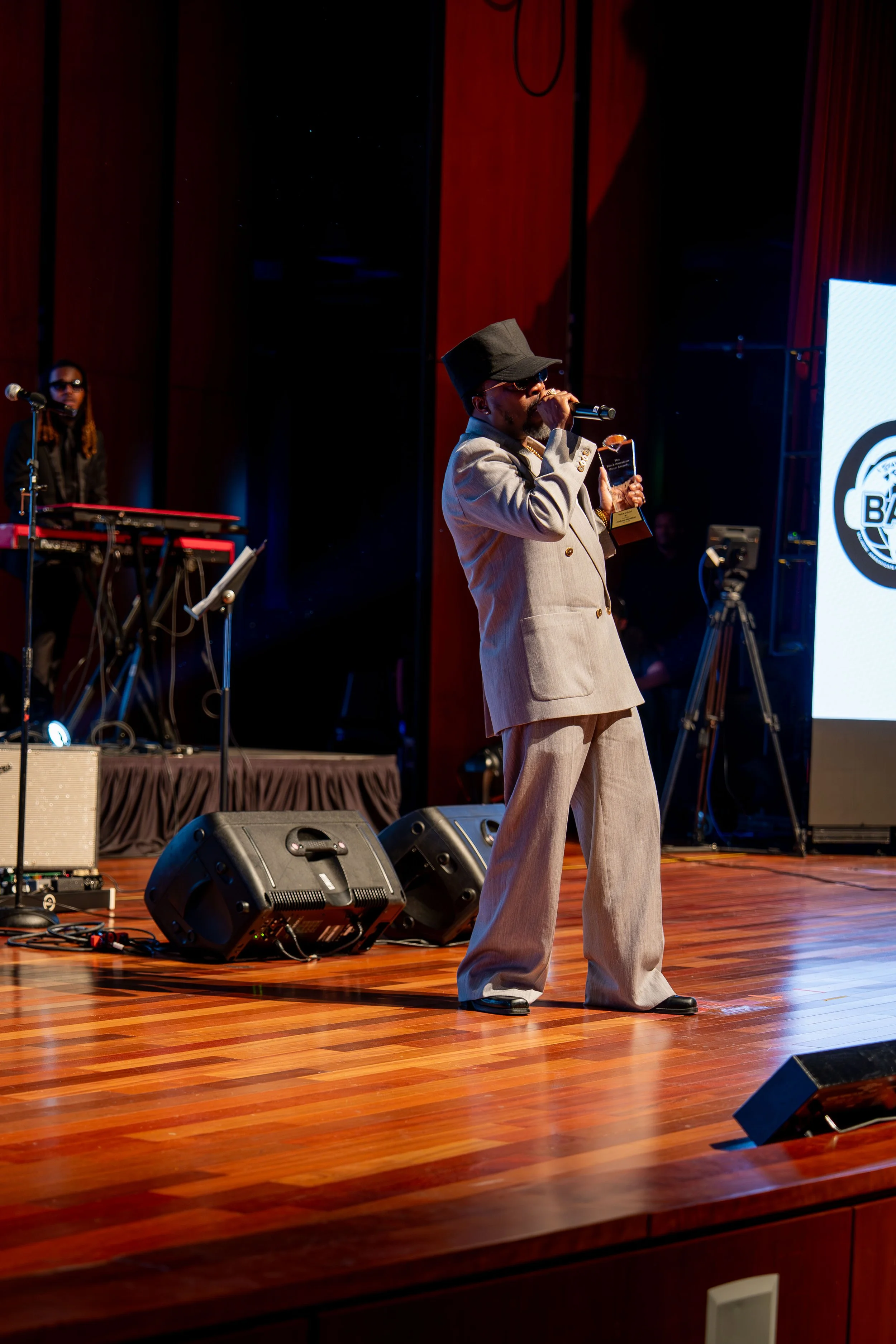 A man wearing a beige suit, black hat, sunglasses, and gold jewelry is singing or speaking into a microphone on a wooden stage. There is a musician with long hair and glasses standing behind a keyboard in the background. The stage has speakers, music equipment, and a large screen with a logo.