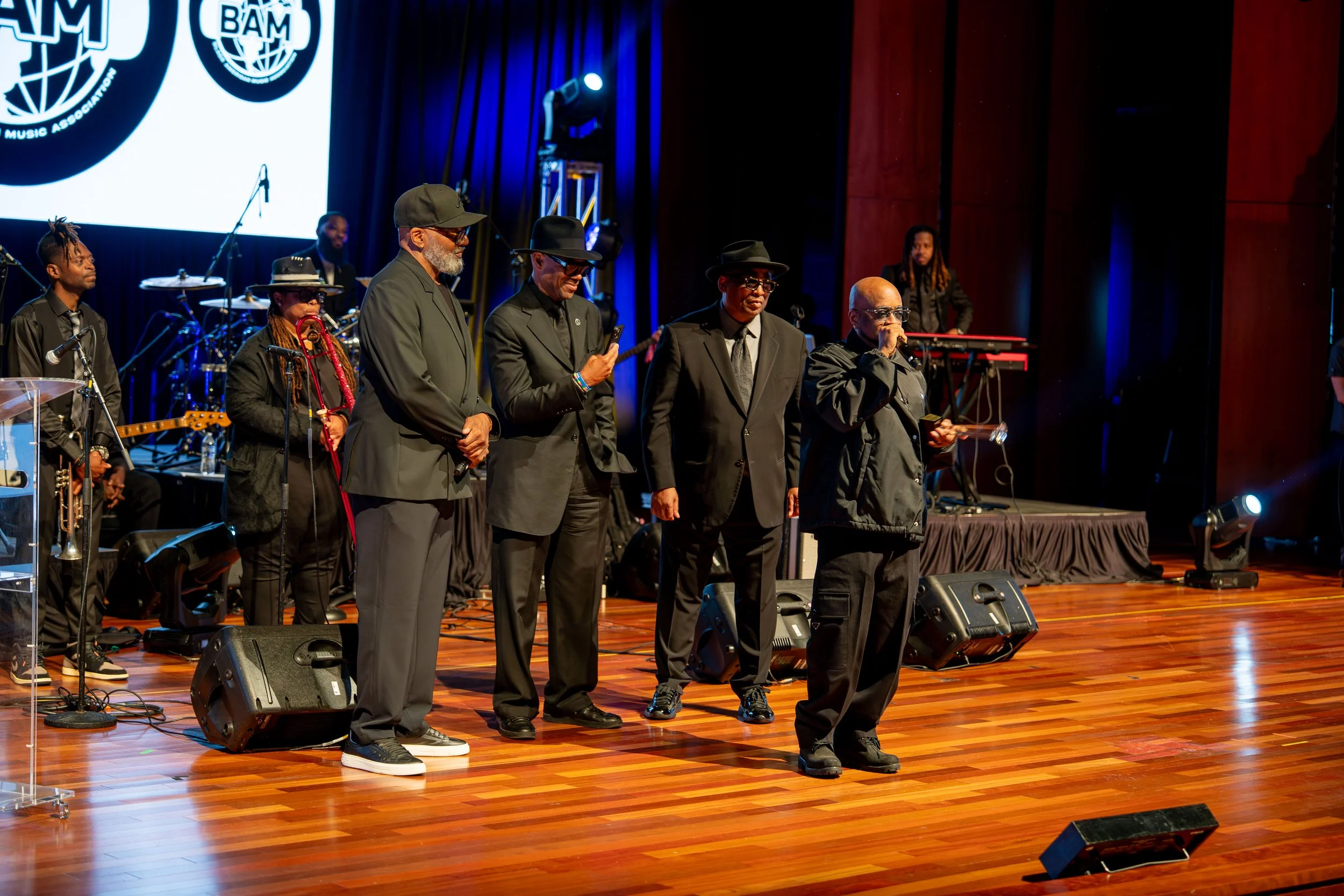A group of seven diverse musicians on stage at a music event, with some holding microphones and others standing near musical instruments, on a wooden stage with a large screen displaying logos in the background.