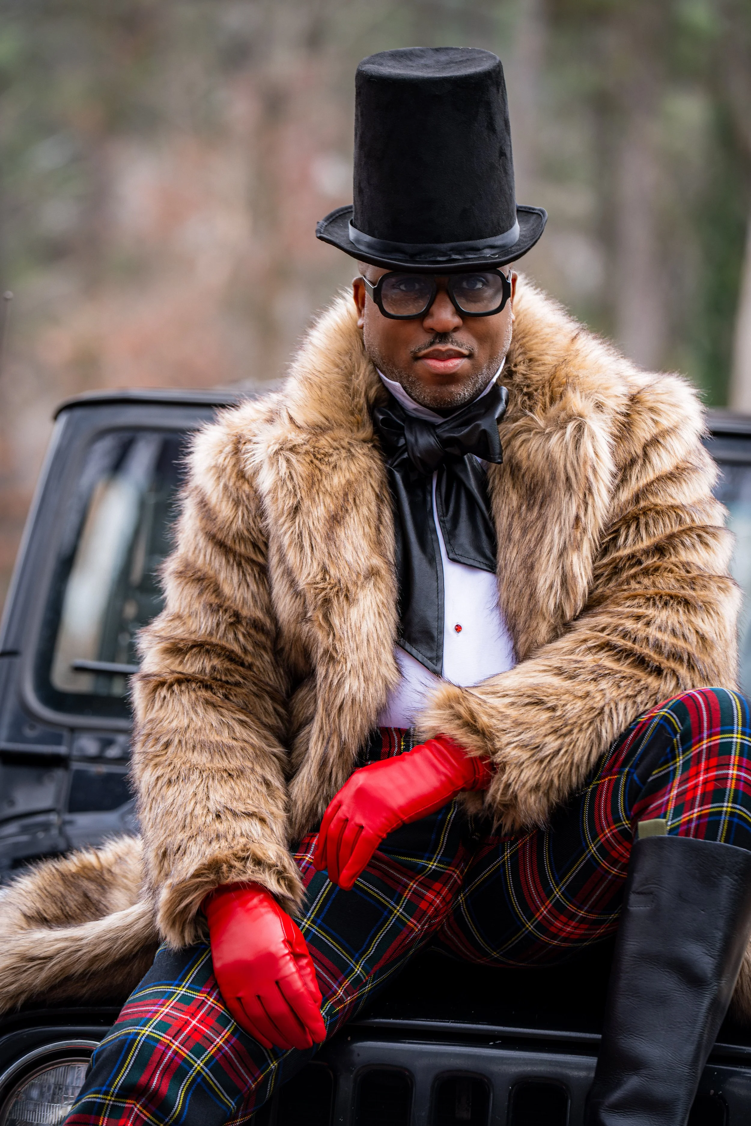 A man dressed in a fur coat, red gloves, and plaid pants, sitting on the hood of a car, wearing large black glasses and a tall black top hat.