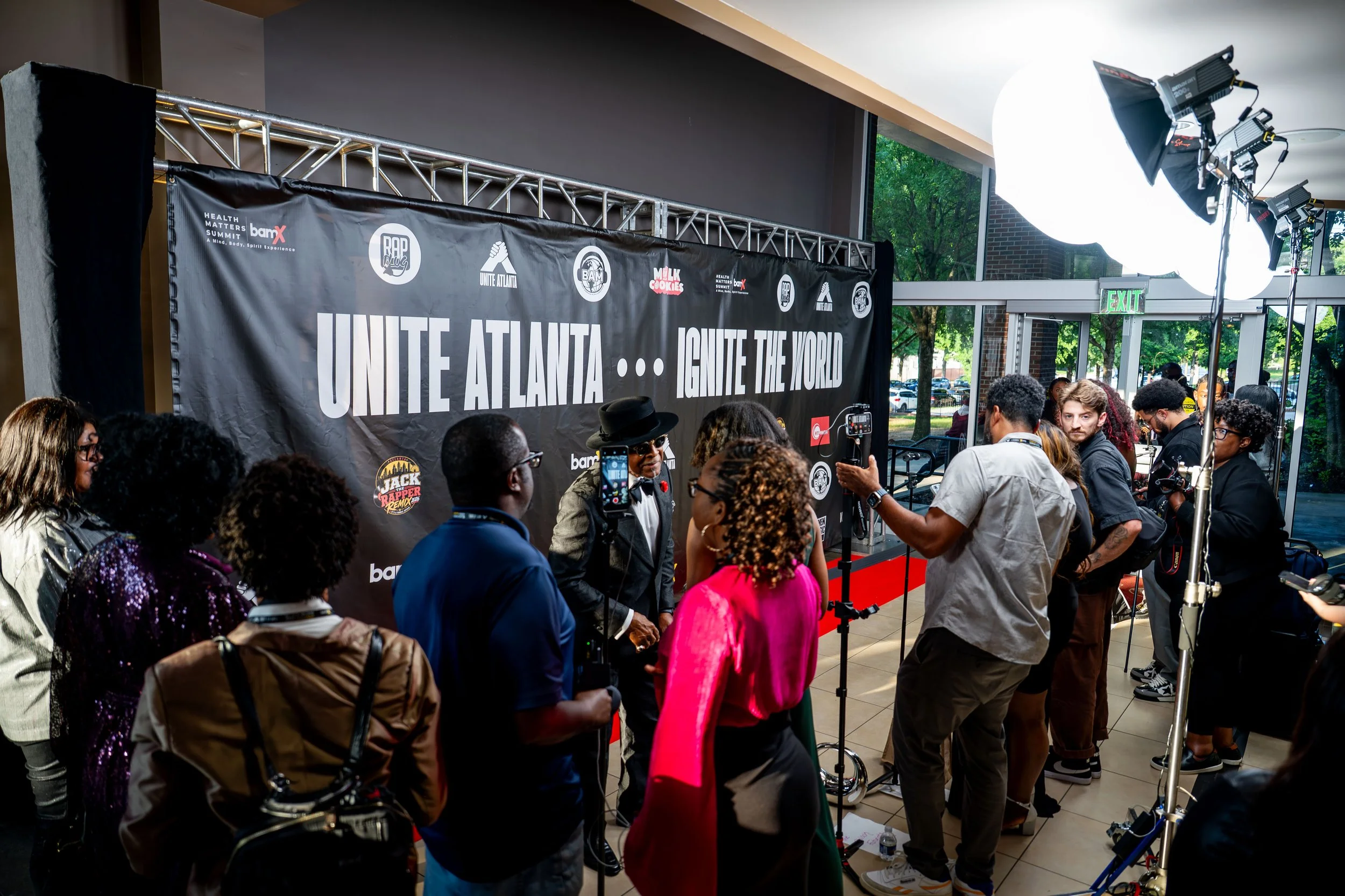 A crowd of people gathered inside a building near a red carpet event, with a large black backdrop reading 'UNITE ATLANTA... IGNITE THE WORLD' and various logos, including health and music themes. Multiple studio lights are set up to illuminate the area.