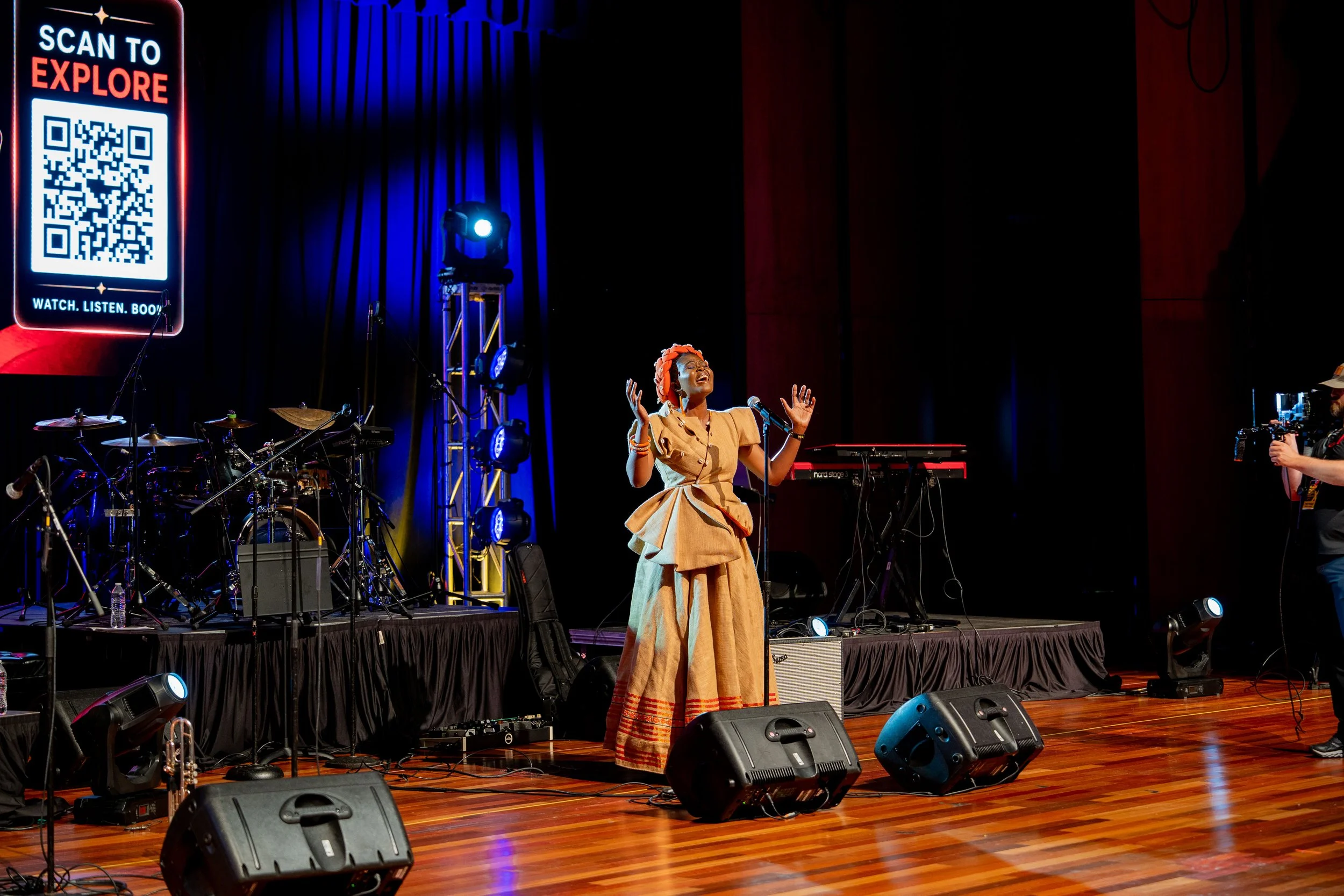 A woman on stage singing with eyes closed, wearing a beige dress and a bright orange headwrap, with musical instruments and stage lighting around her.