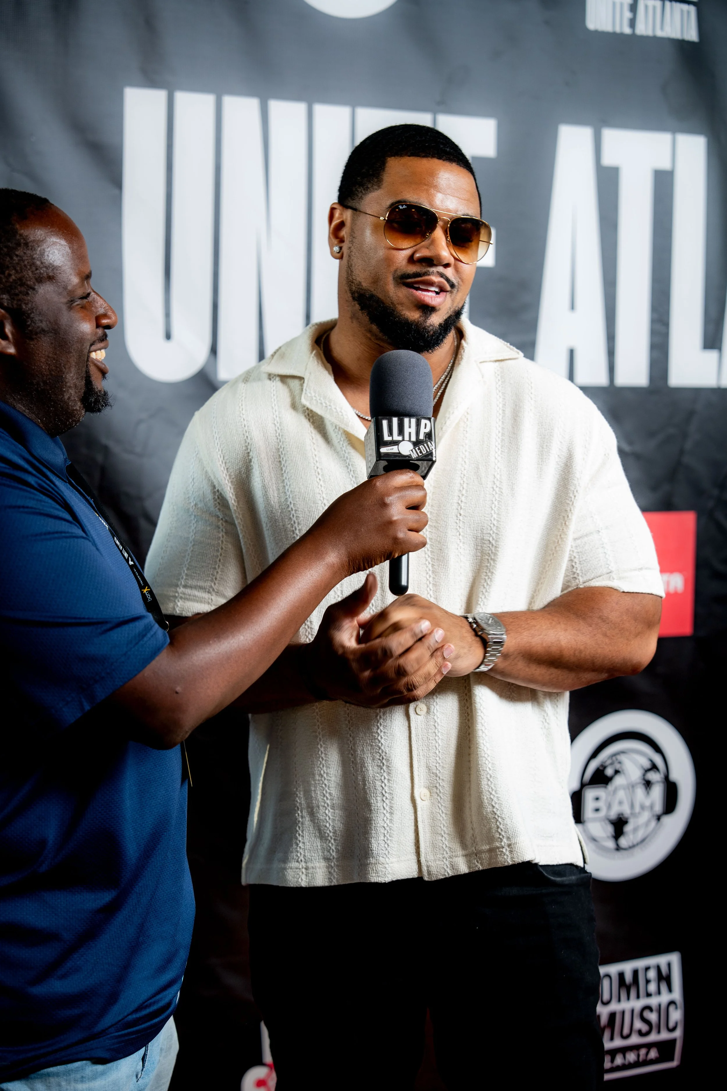 Close-up of two men, one wearing sunglasses, engaged in conversation in front of a backdrop with text, at an indoor event.