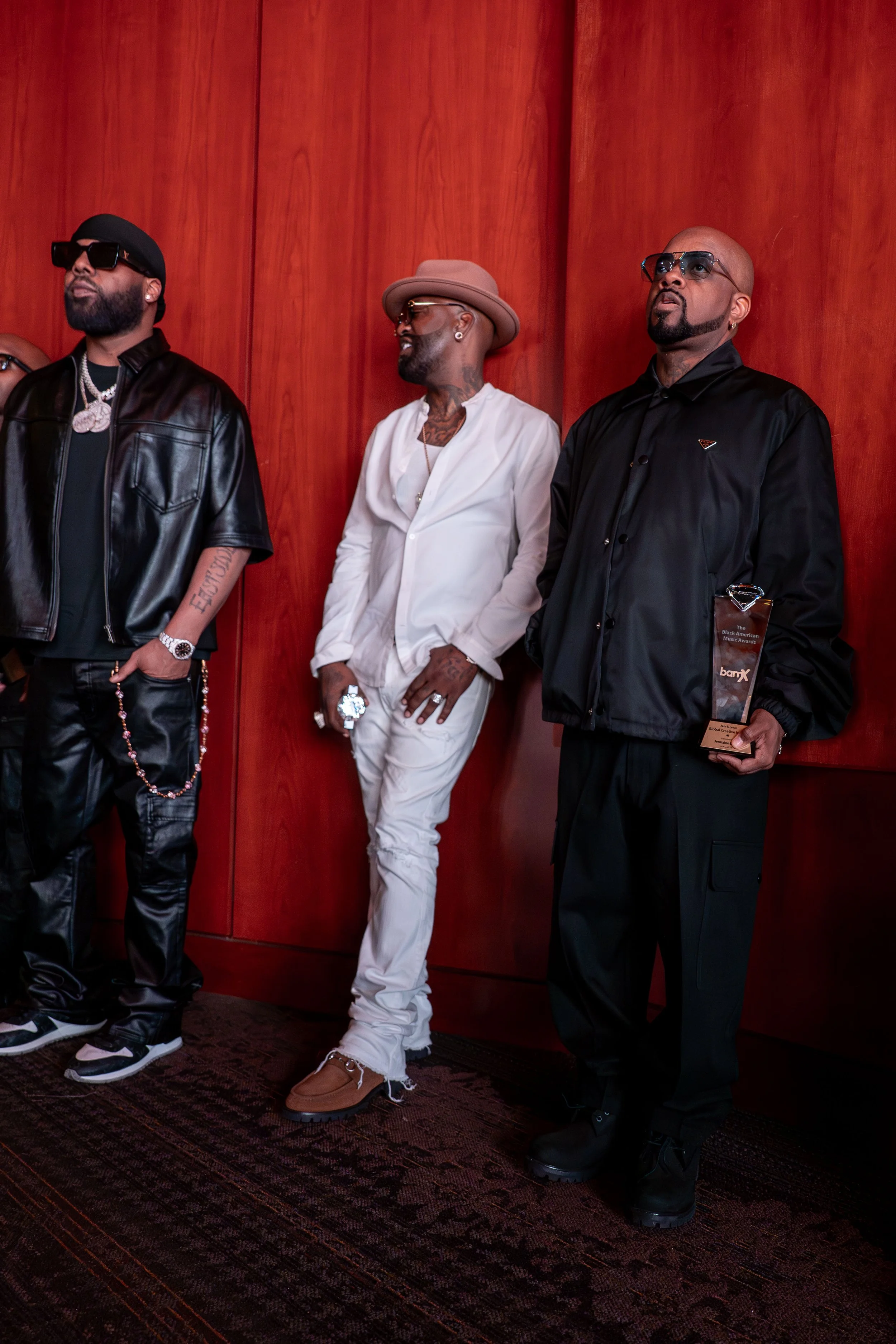 Three men in stylish clothing standing against a red wooden panel wall.