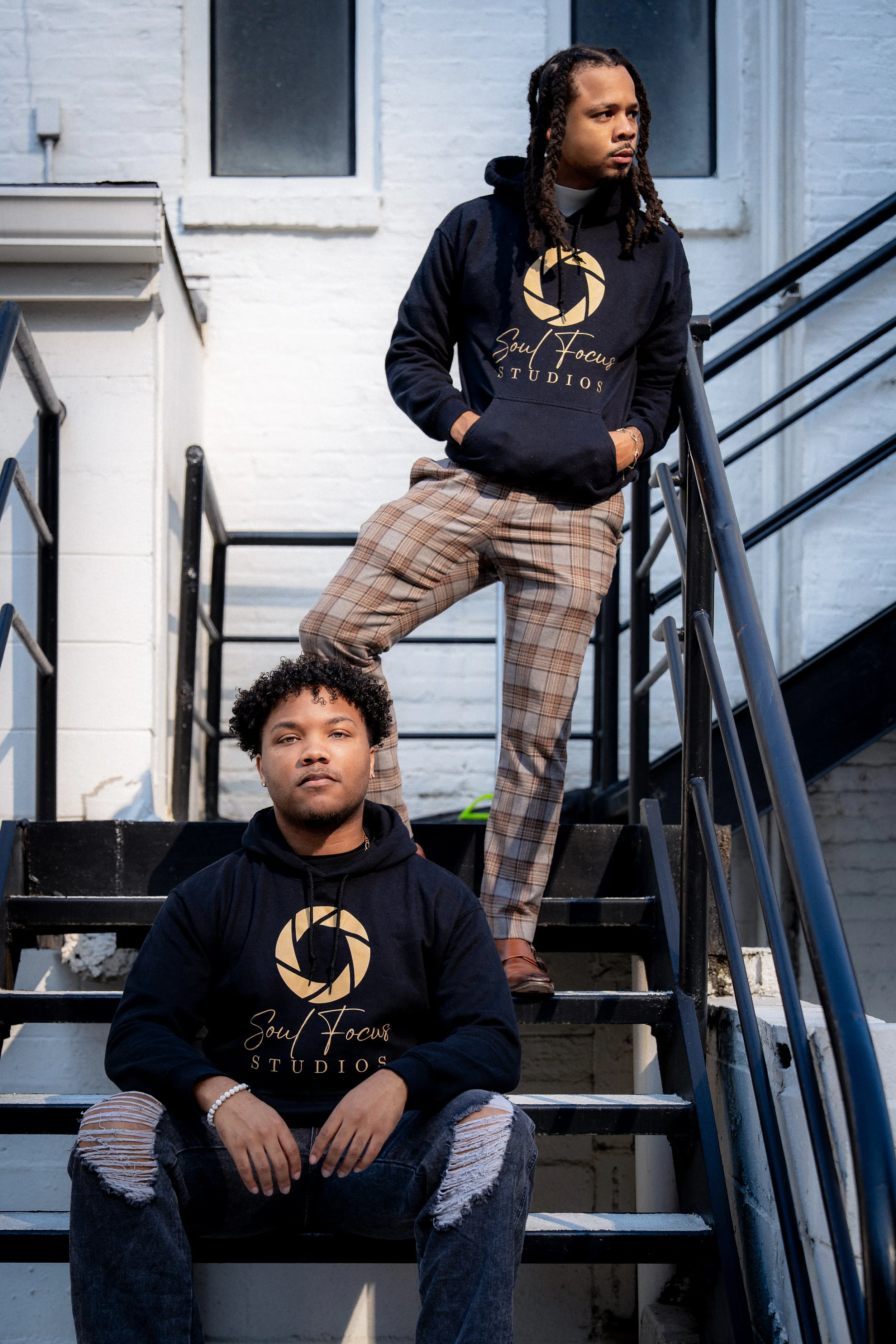 Two young men sitting on outdoor staircase, both wearing black hoodies with gold logo and text that says "Soul Focus Studios." The man in the foreground has curly hair, ripped jeans, and is looking directly at the camera. The man in the background has dreadlocks, plaid pants, and is looking to the side.