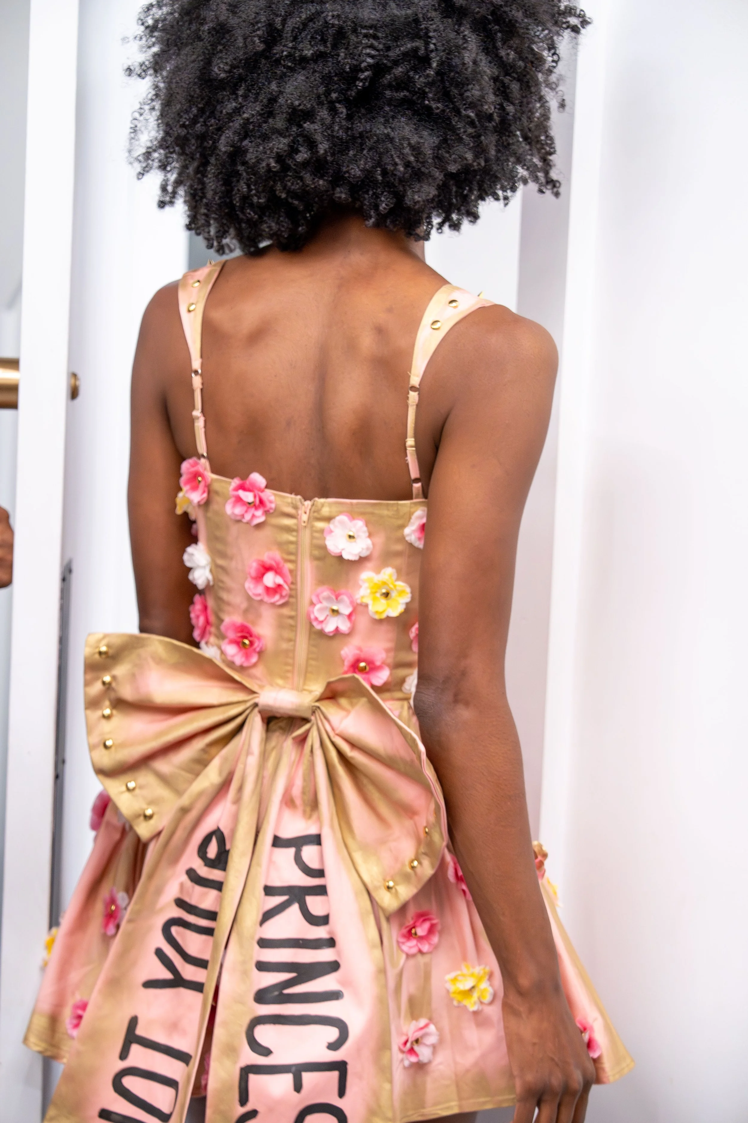 Back of a woman with curly black hair wearing a pink dress with floral embroidery and a large bow, standing in front of a white wall.