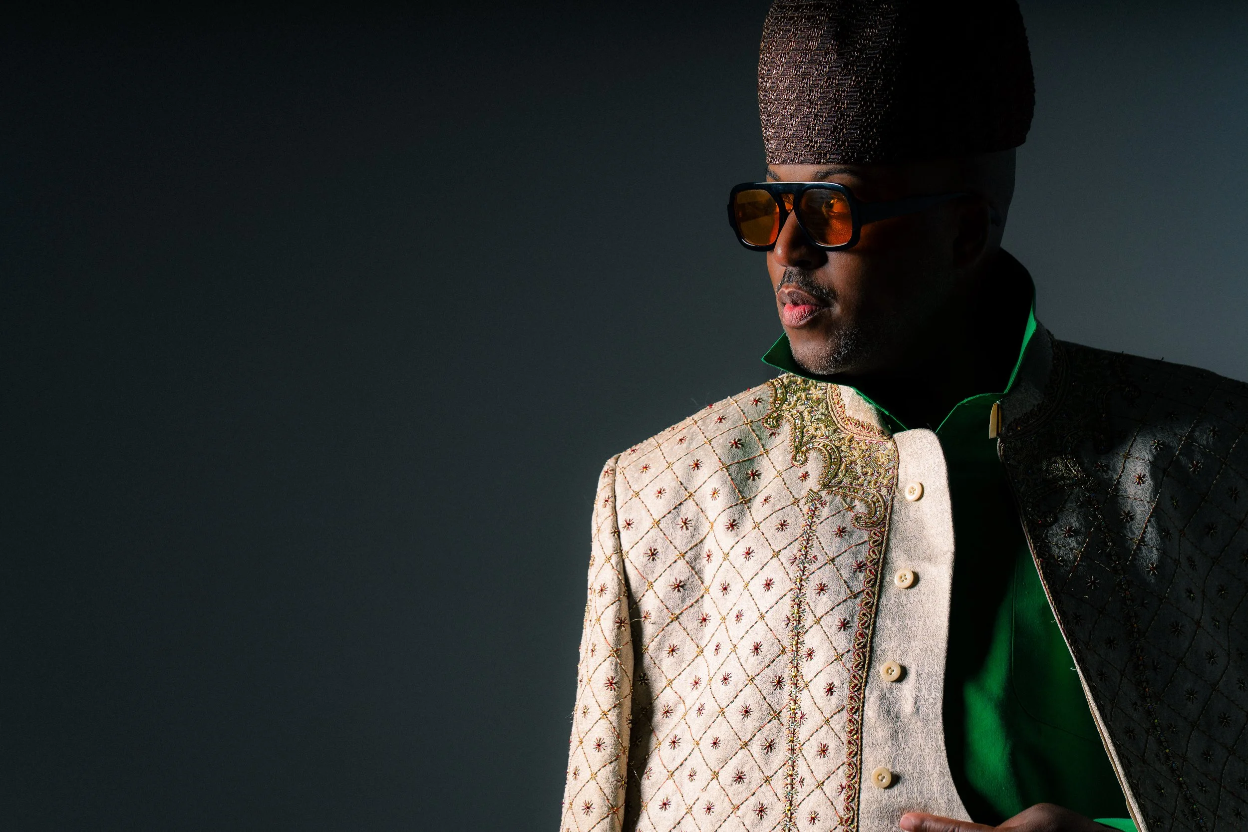 A man wearing a traditional African outfit with intricate embroidery, large sunglasses with orange lenses, and a tall, textured hat. The background is dark and plain.