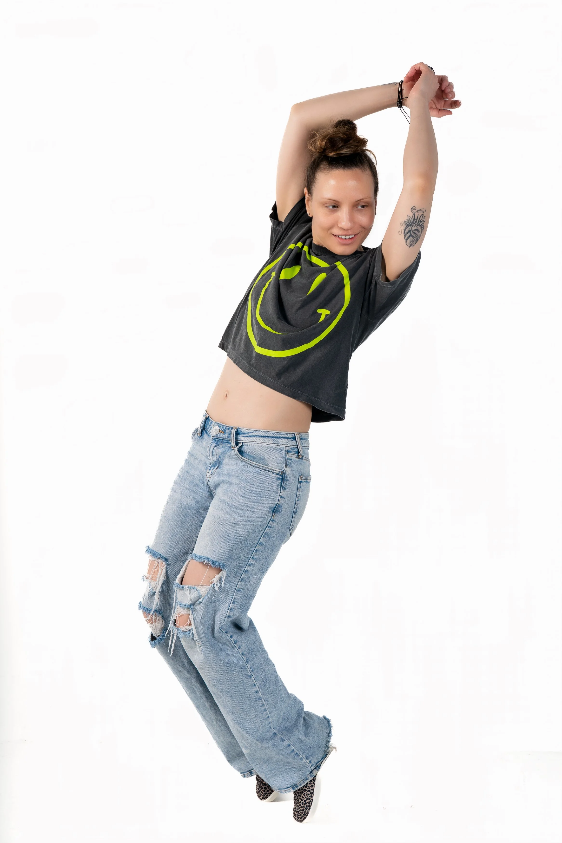 A woman stretching with her arms above her head, wearing a black T-shirt with a yellow smiley face, ripped jeans, and leopard print shoes, standing against a plain white background.
