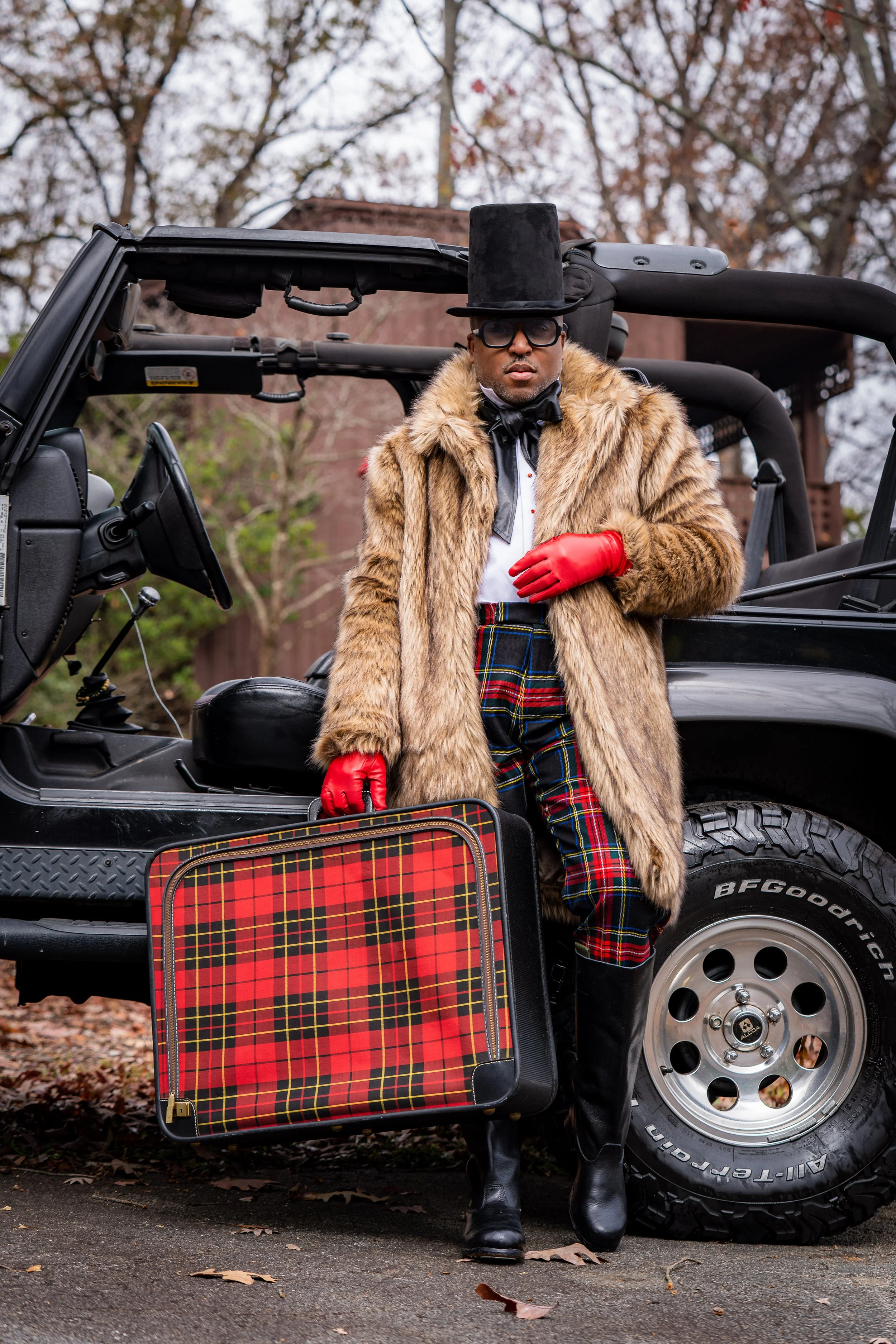 Person dressed in vintage-style clothing, including a tall black top hat, fur coat, red gloves, and plaid pants, standing next to a black off-road vehicle with an open door. The person is holding a plaid suitcase, and the background features leafless trees and an overcast sky.