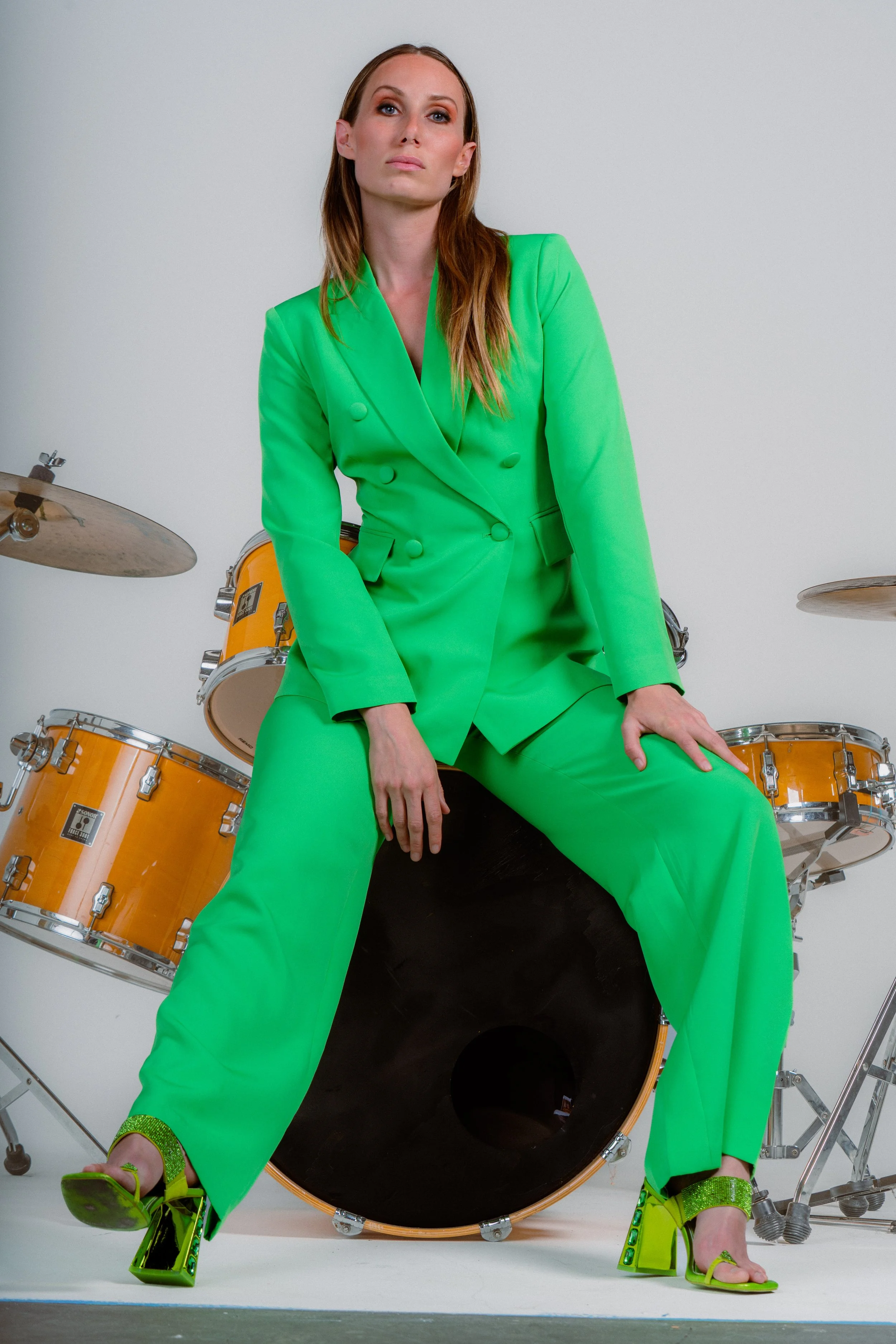 A woman in a bright green suit and matching green high-heeled shoes sitting on a large black drum, with drums in the background.