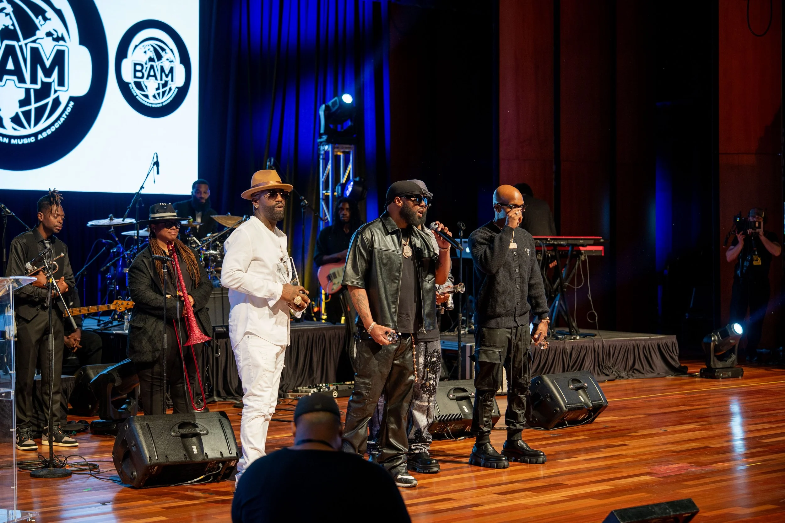A group of musicians and singers perform on stage with blue lighting and a large screen displaying the BAM logo in the background.