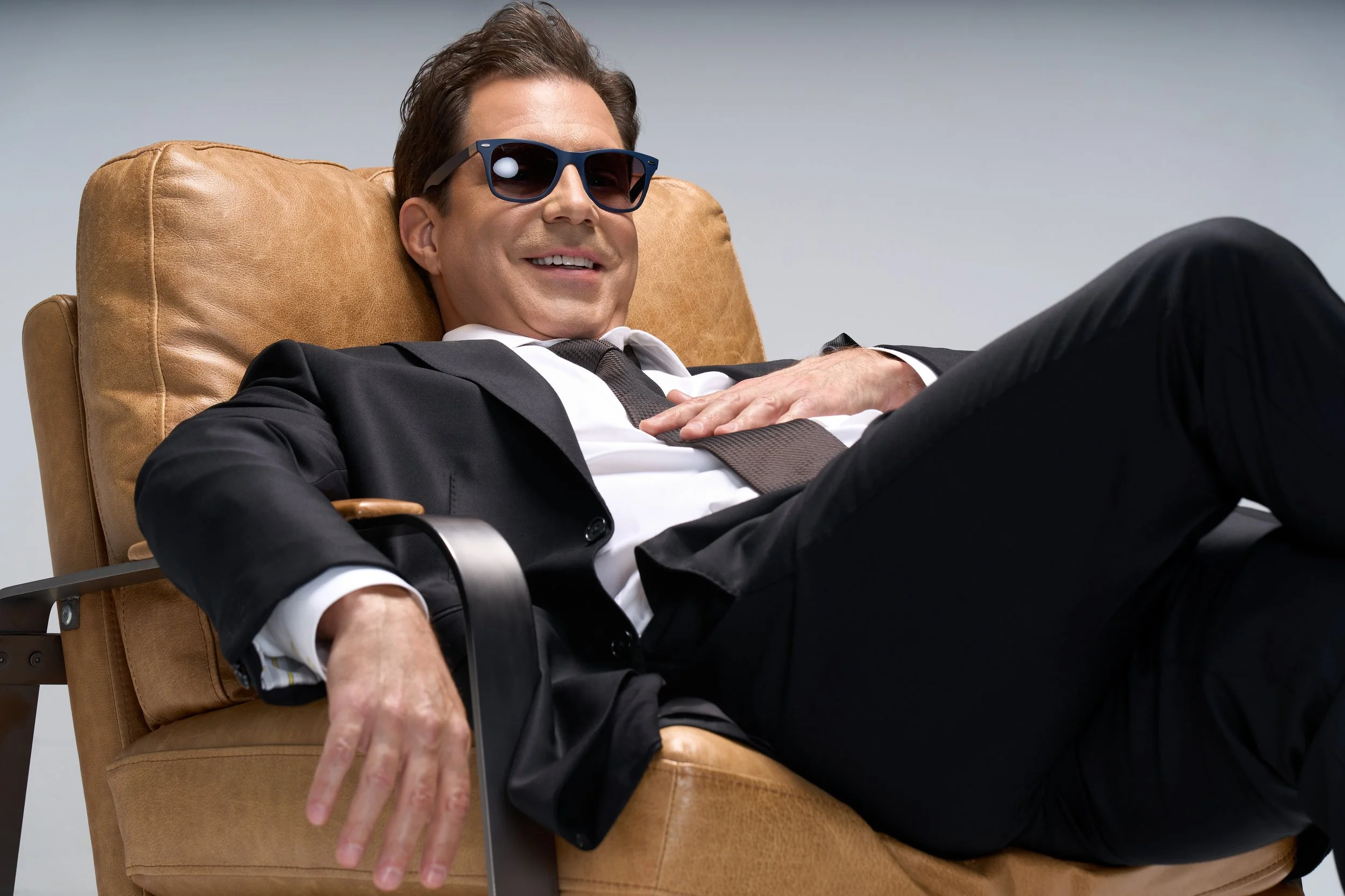 A man in a black suit, white shirt, and sunglasses lounging in a tan leather chair with a relaxed smile.