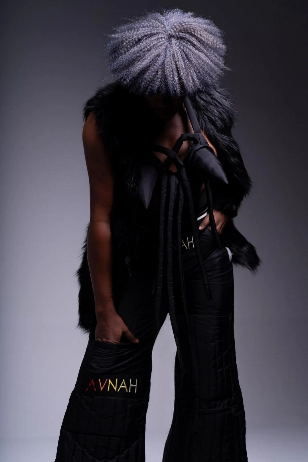 A person wearing a lavender wig, black fur vest, and black pants with colorful text, posing against a gray background.