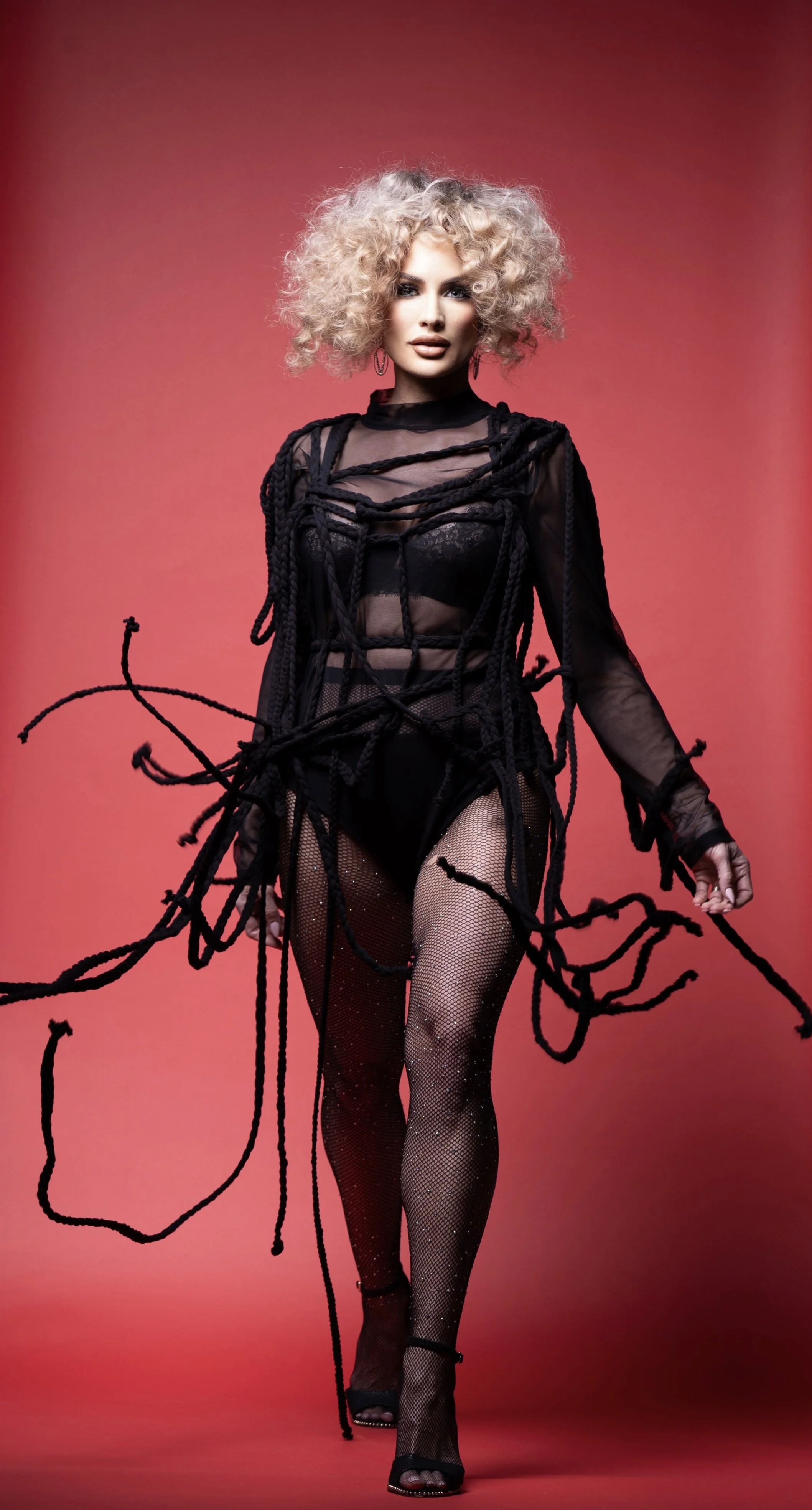A woman with large, curly blonde hair wearing sheer black clothing, fishnet stockings, high heels, and black rope fashion details, standing against a pink gradient background.