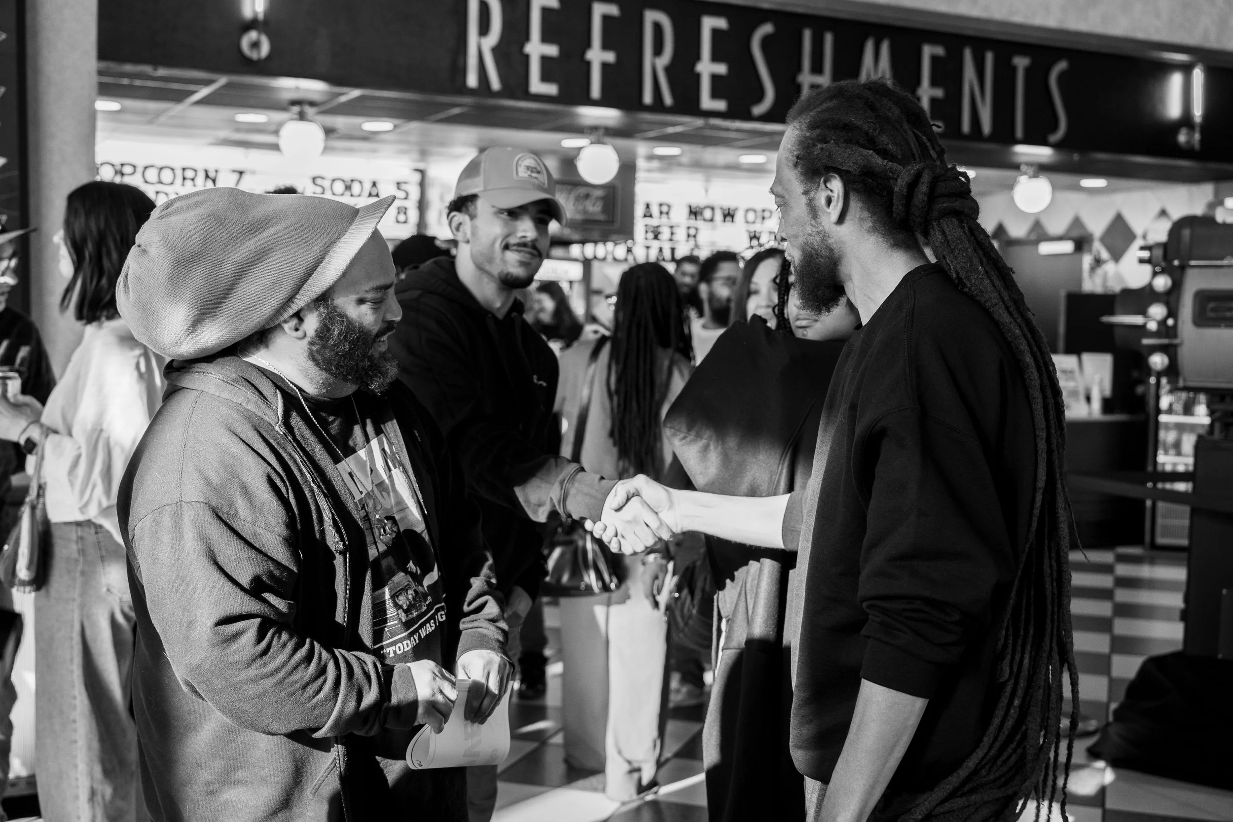 Two men shaking hands in a crowded indoor setting, with a sign in the background that reads 'REFRESHMENTS.' The man on the left is wearing a hooded jacket and a hat, while the man on the right has long dreadlocks and is dressed in a dark shirt. They are smiling and engaging in a friendly greeting.
