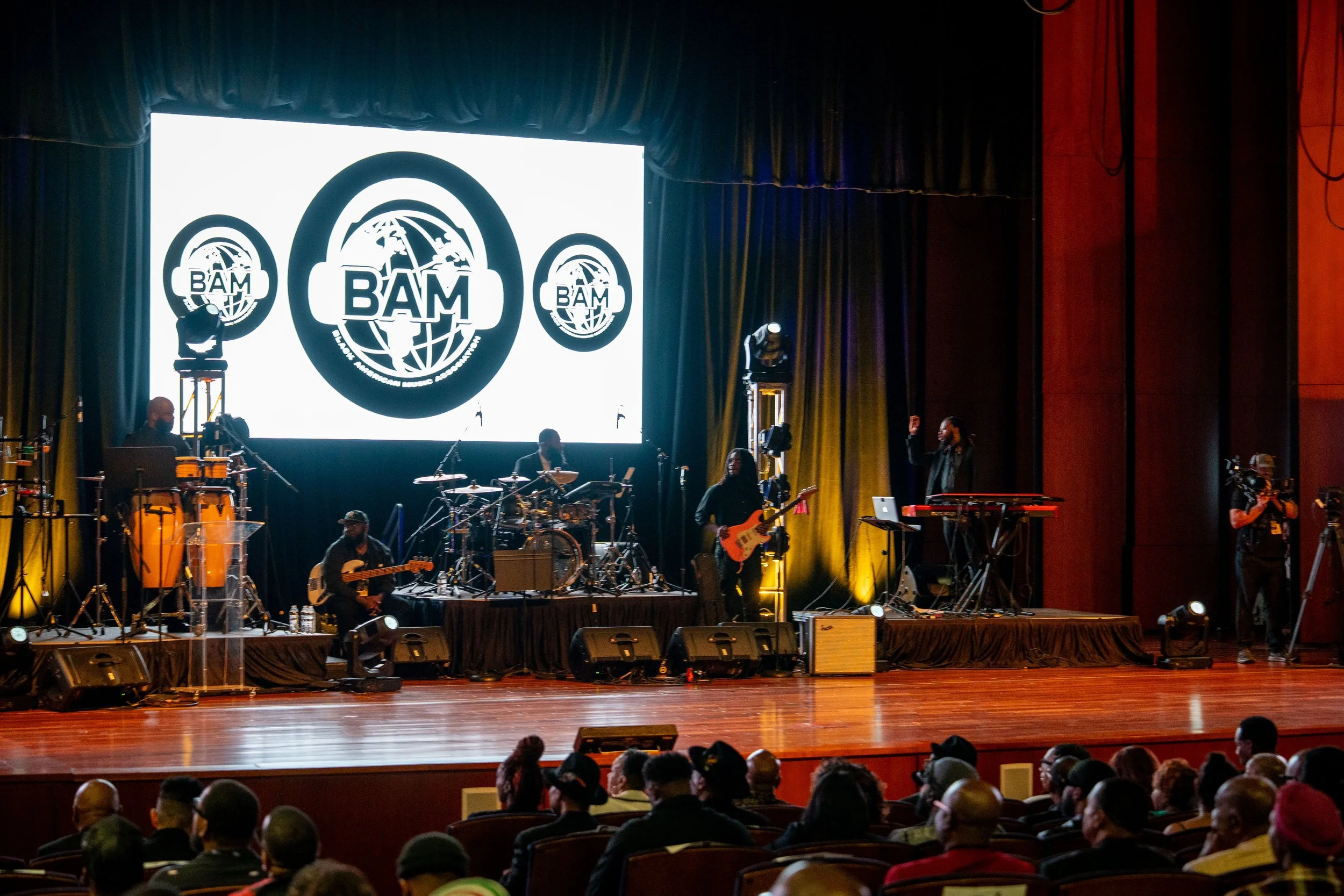 A stage with musical instruments and a large screen displaying the logo 'BAM,' surrounded by an audience in a concert hall.