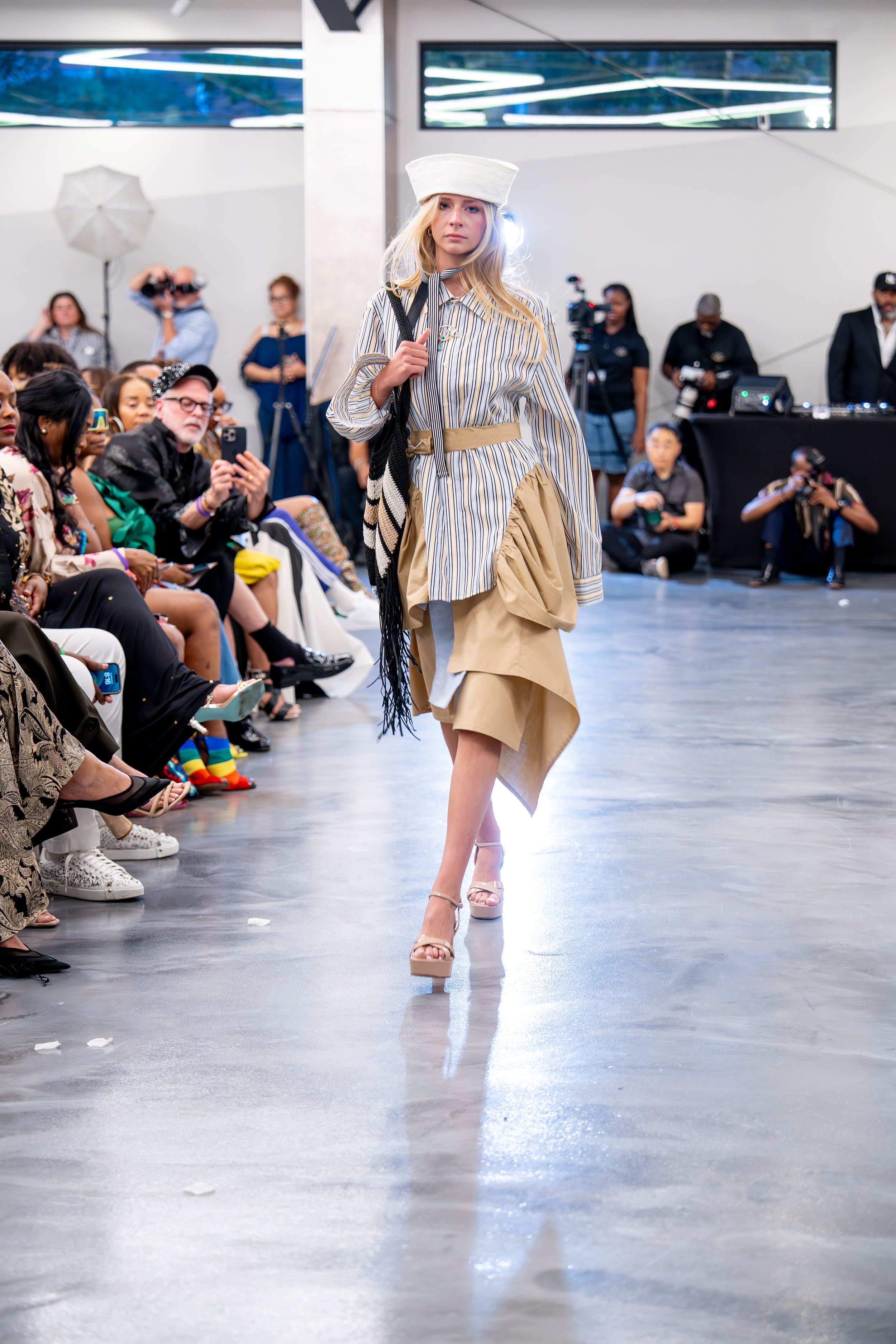 A female model walking on a runway during a fashion show, surrounded by spectators and photographers, with a modern indoor setting and bright lighting.