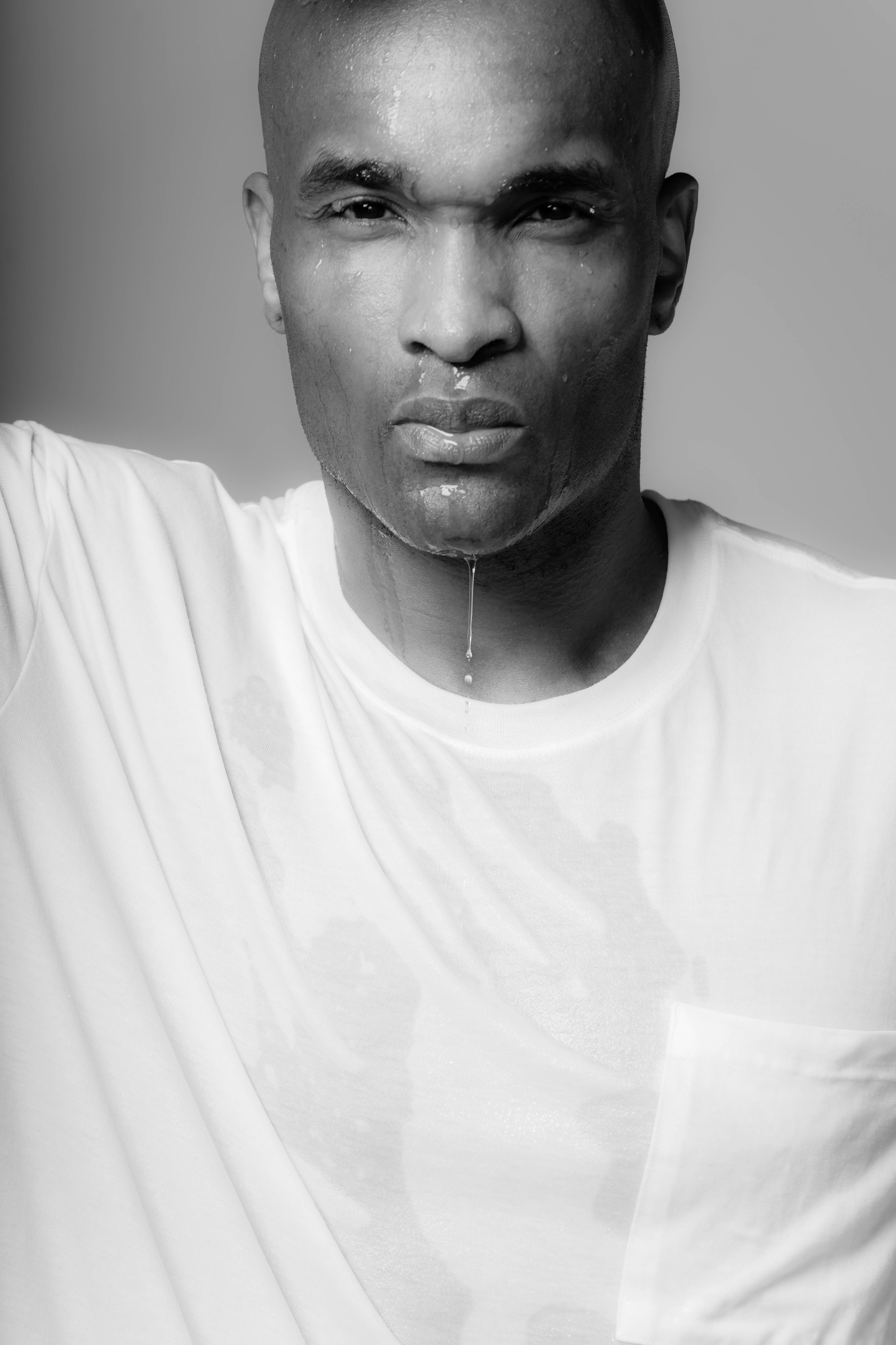 A black and white portrait of a man with water on his face and shoulders, wearing a white shirt, against a plain background.
