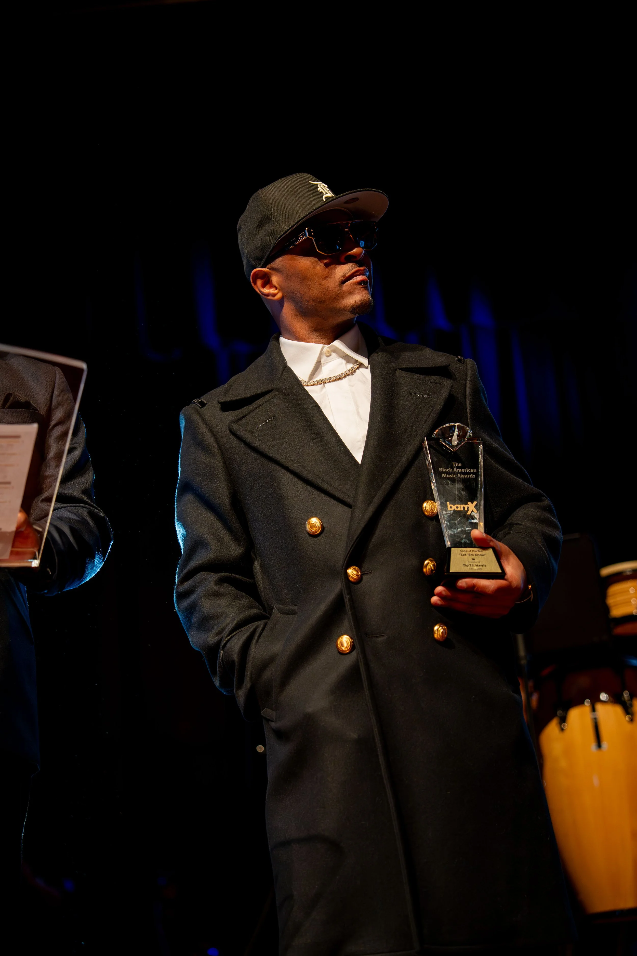 Man wearing black cap and sunglasses, dressed in a black coat with gold buttons, holding a clear award trophy at an event, standing in front of a dark blue background with stage lighting.