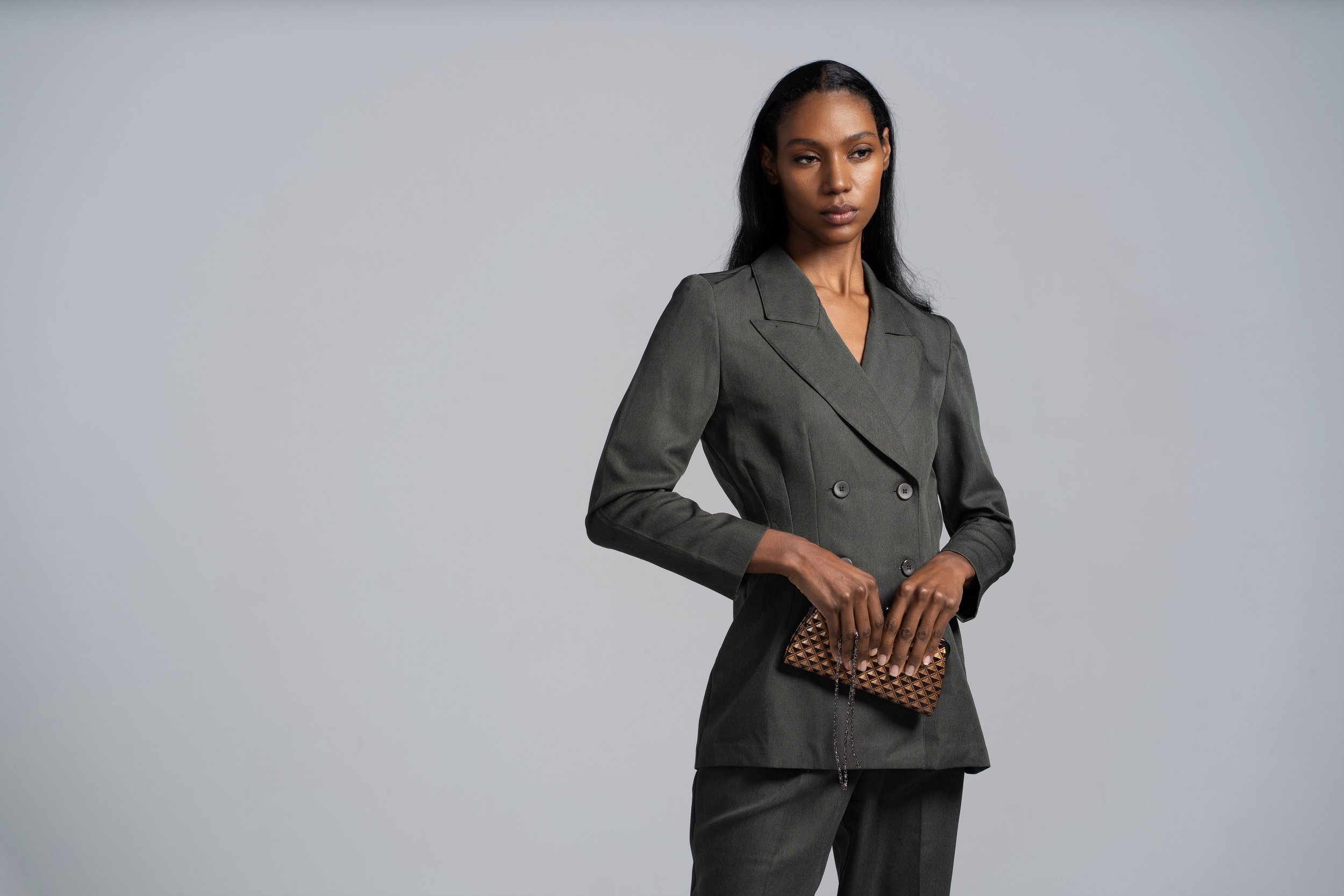 A woman with dark hair wearing a tailored grey blazer and matching skirt, holding a brown patterned clutch purse, standing against a plain light grey background.