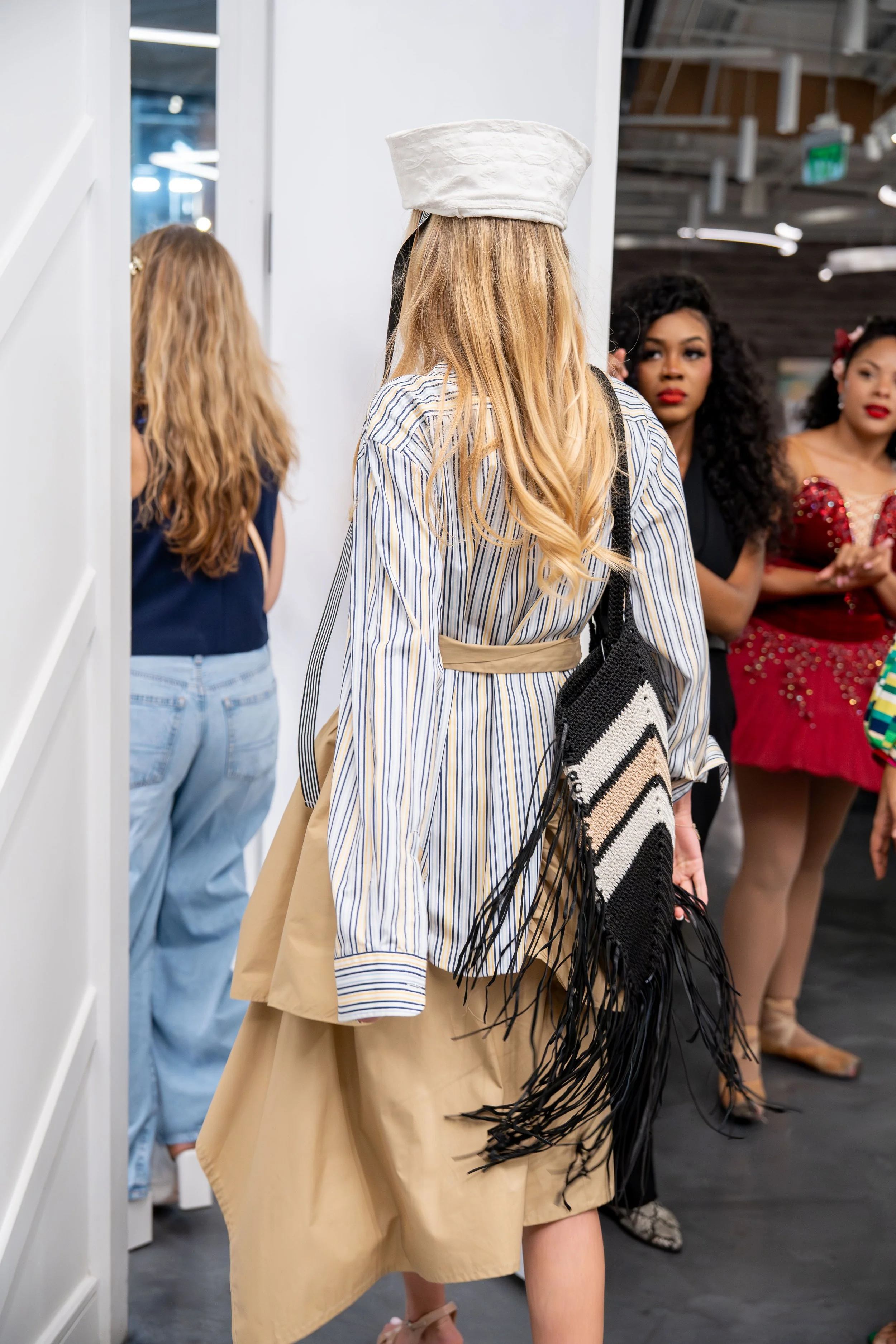 People standing in line at an indoor event, with three women visible, dressed in fashionable clothing, including a woman with long blonde hair wearing a striped shirt and beige skirt, and two women in red and black outfits.
