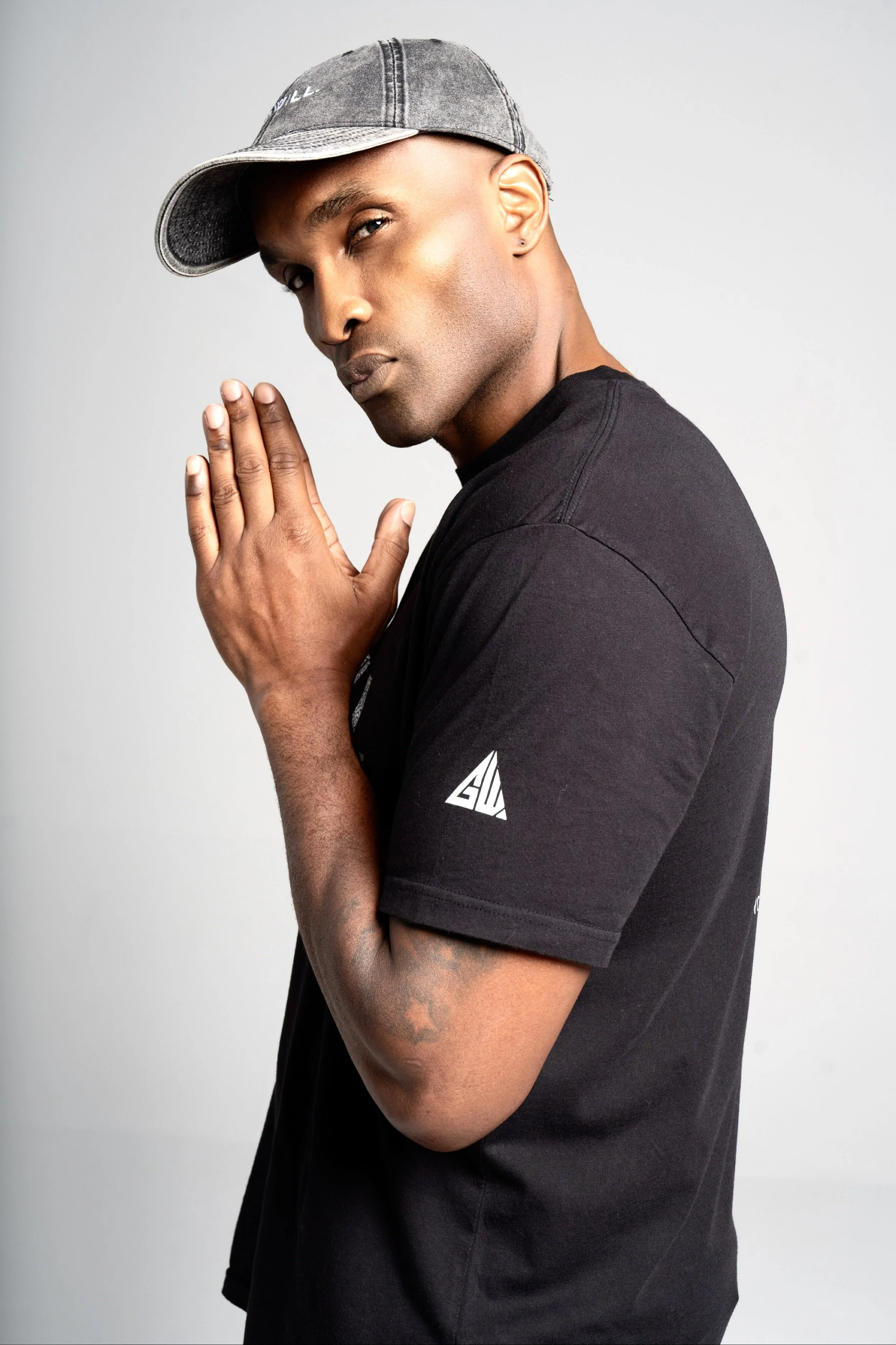 A man with light brown skin and a tattoo on his forearm, wearing a gray baseball cap tilted to the side and a black T-shirt with a white logo on the sleeve. He is posing with his hands pressed together near his face and looking at the camera against a plain white background.