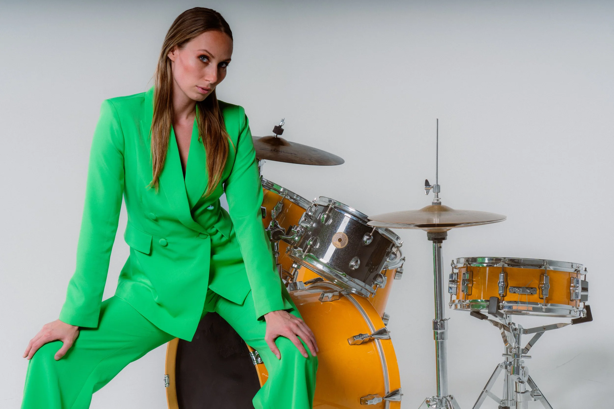 A woman in a bright green suit sitting on a black drum, next to a regular drum set with yellow-orange drums.