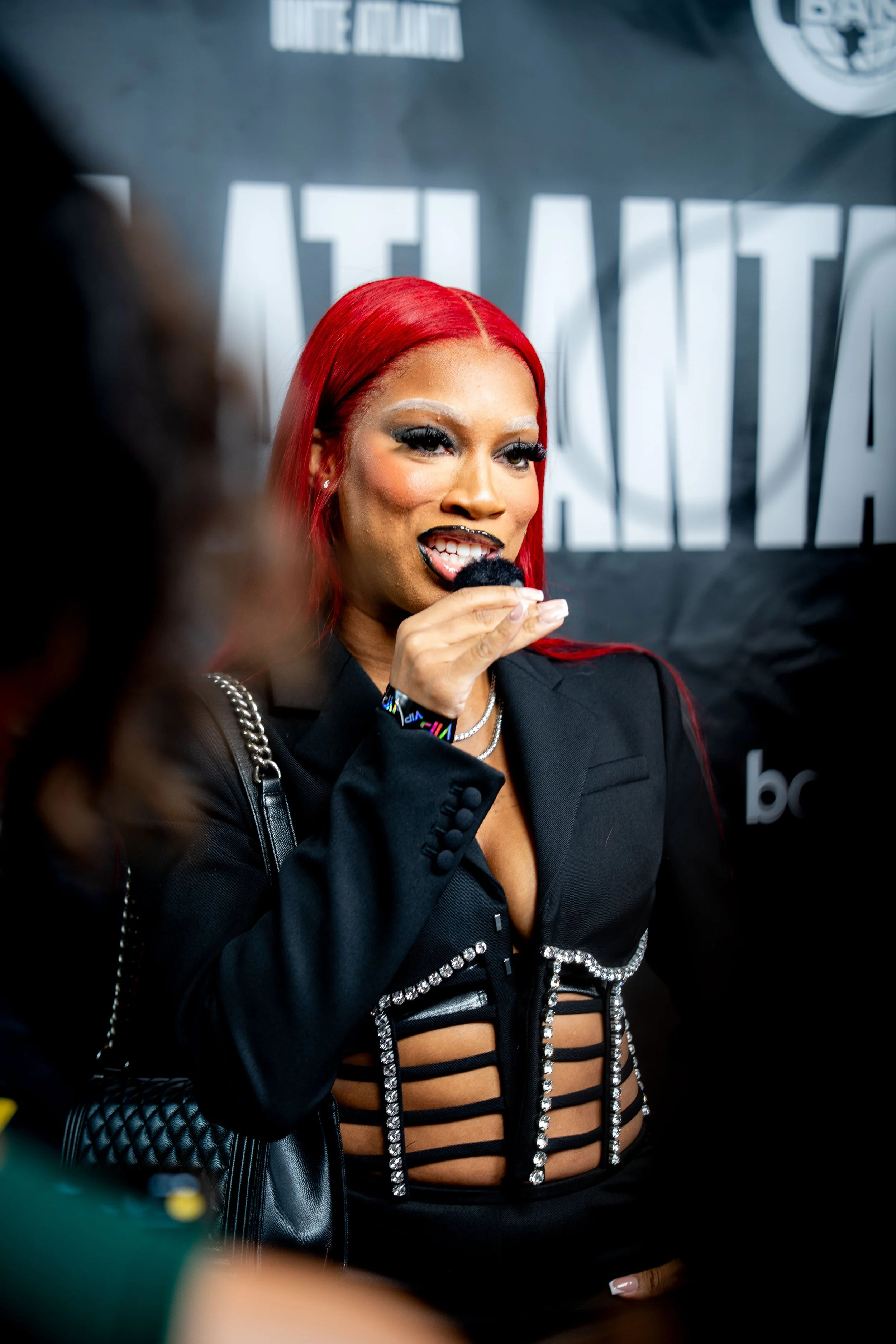 A woman with bright red hair speaking into a microphone at an event. She is wearing a black outfit with metallic accents and has theatrical makeup.