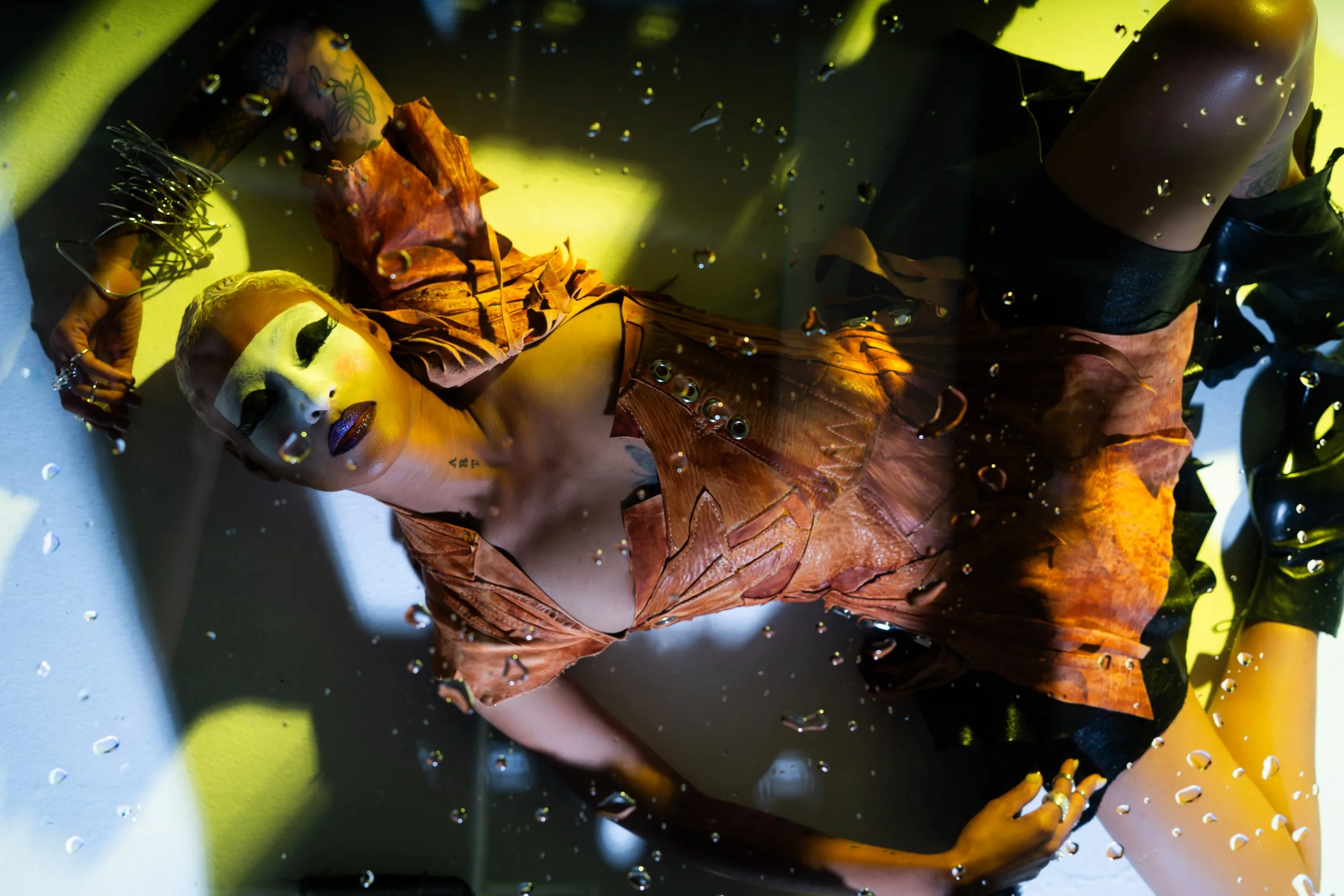A woman with platinum blonde hair and dramatic makeup lying in water with yellow lighting. She is wearing a brown leather-style jacket and has black nail polish and jewelry. Water droplets are visible around her.