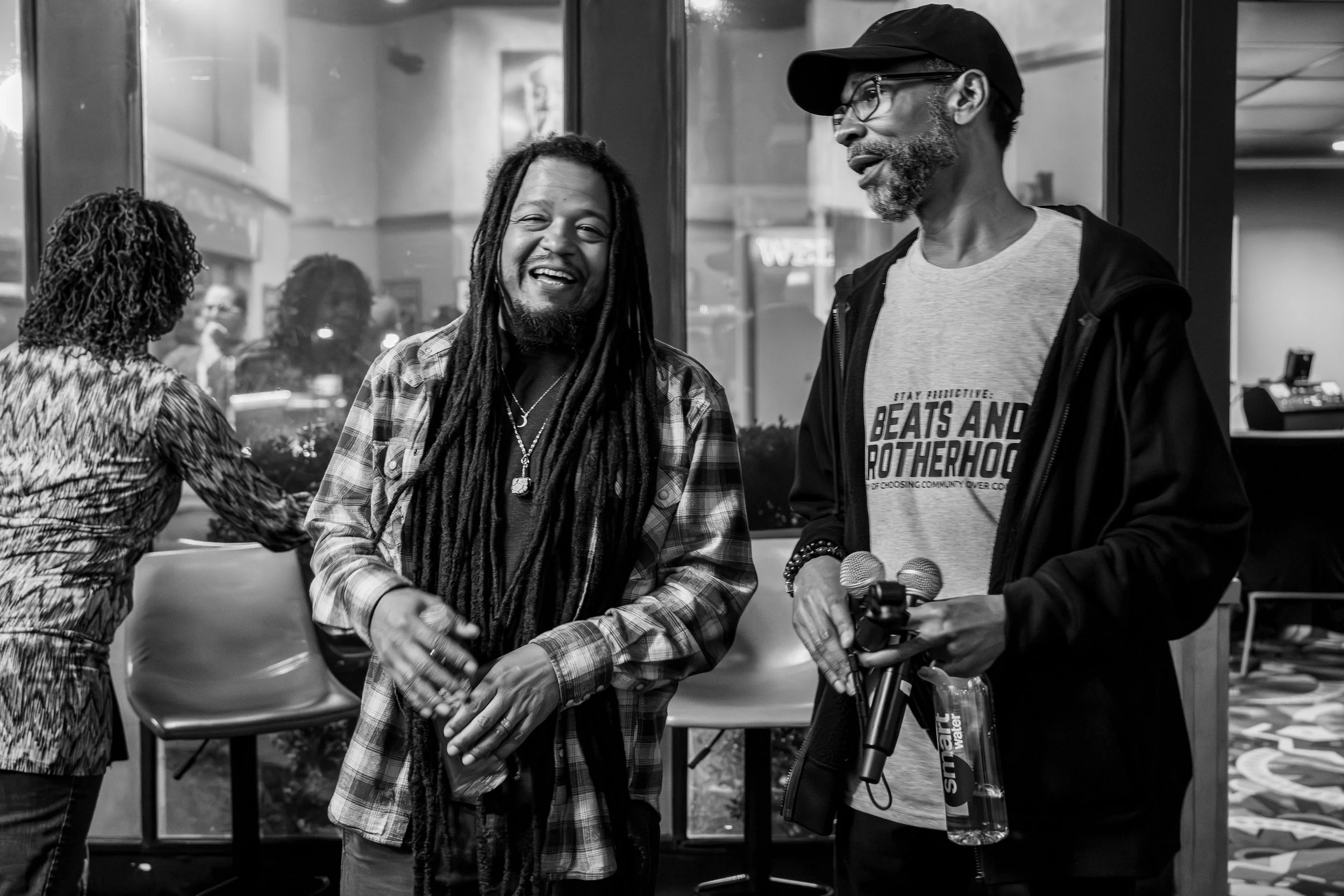 Two men shaking hands and smiling inside a store or cafe, with other people in the background. One man has dreadlocks, the other wears glasses and a cap.