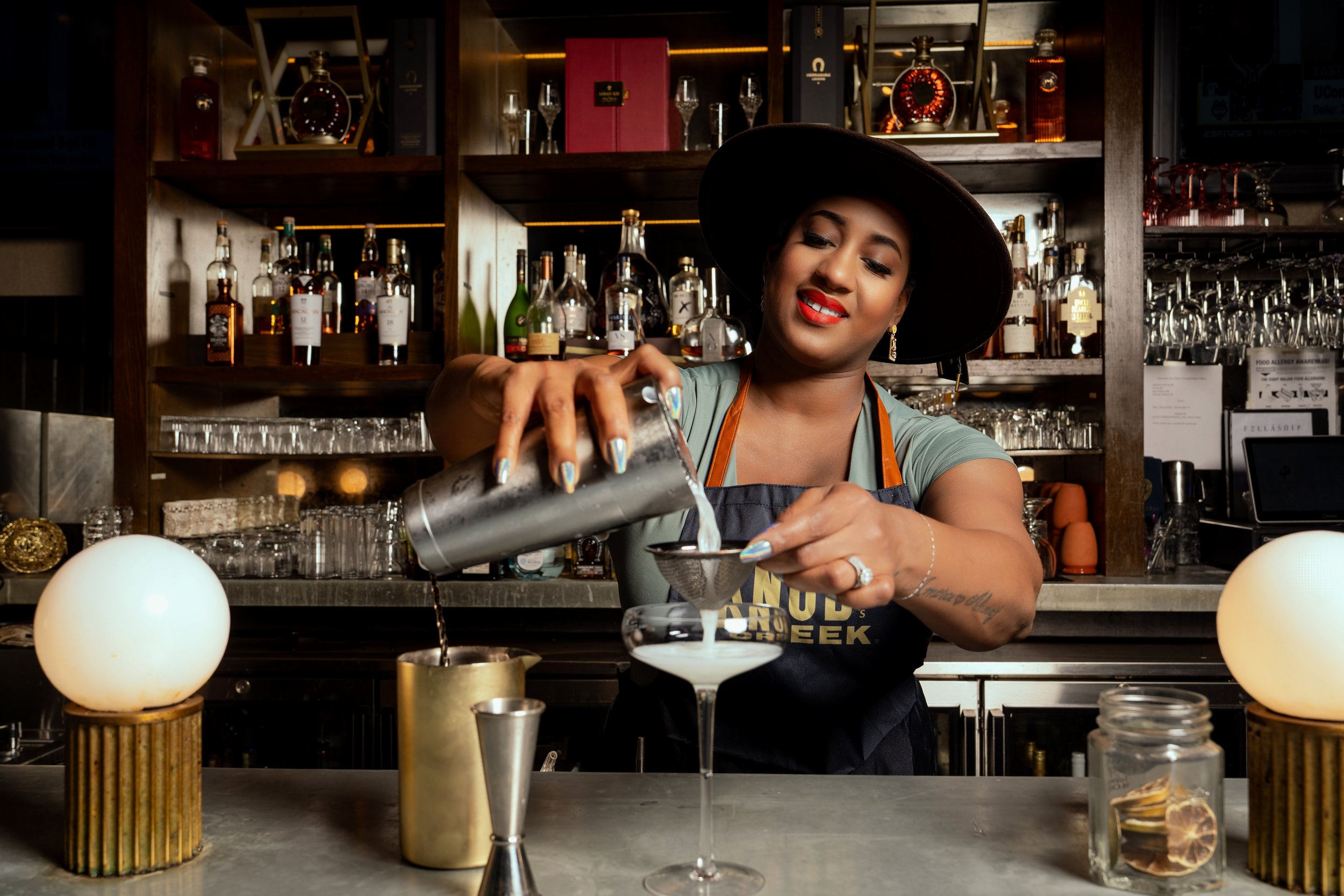 A woman with dark hair, red lipstick, and earrings is smiling while pouring a cocktail through a strainer into a glass at a bar. She is wearing a wide-brimmed hat and an apron. The bar has bottles of alcohol, glasses, and decorative lighting in the background.