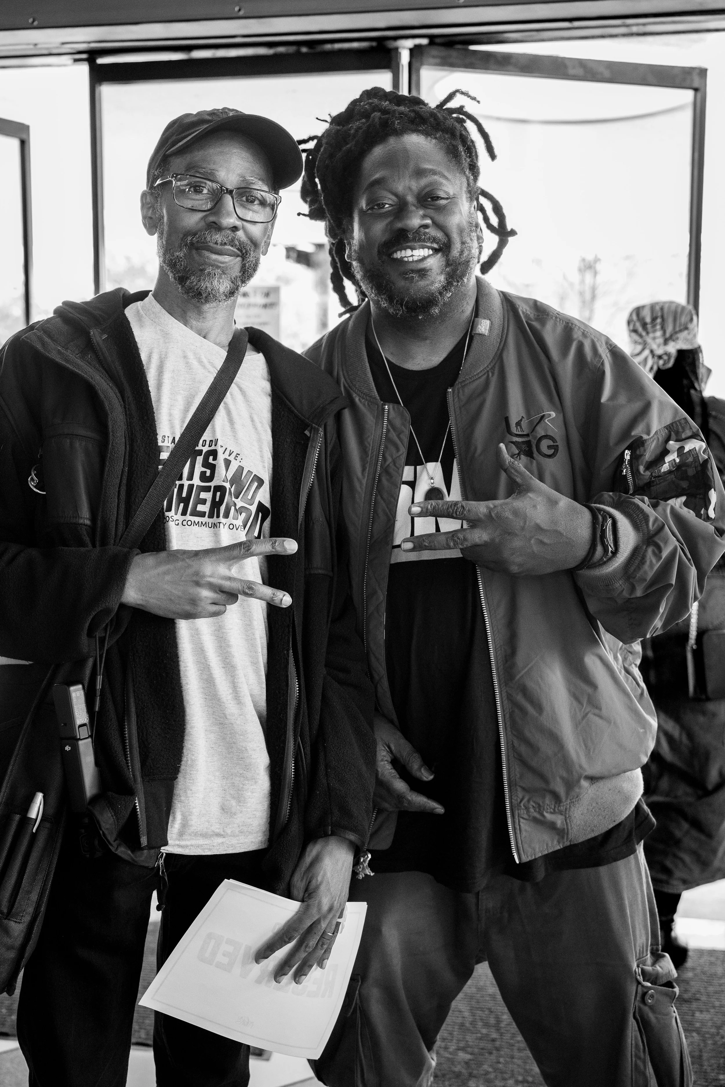 Two men smiling and posing together inside a building, making peace signs with their hands. One man is wearing glasses, a cap, and a sweatshirt, while the other has dreadlocks and a jacket with a logo.