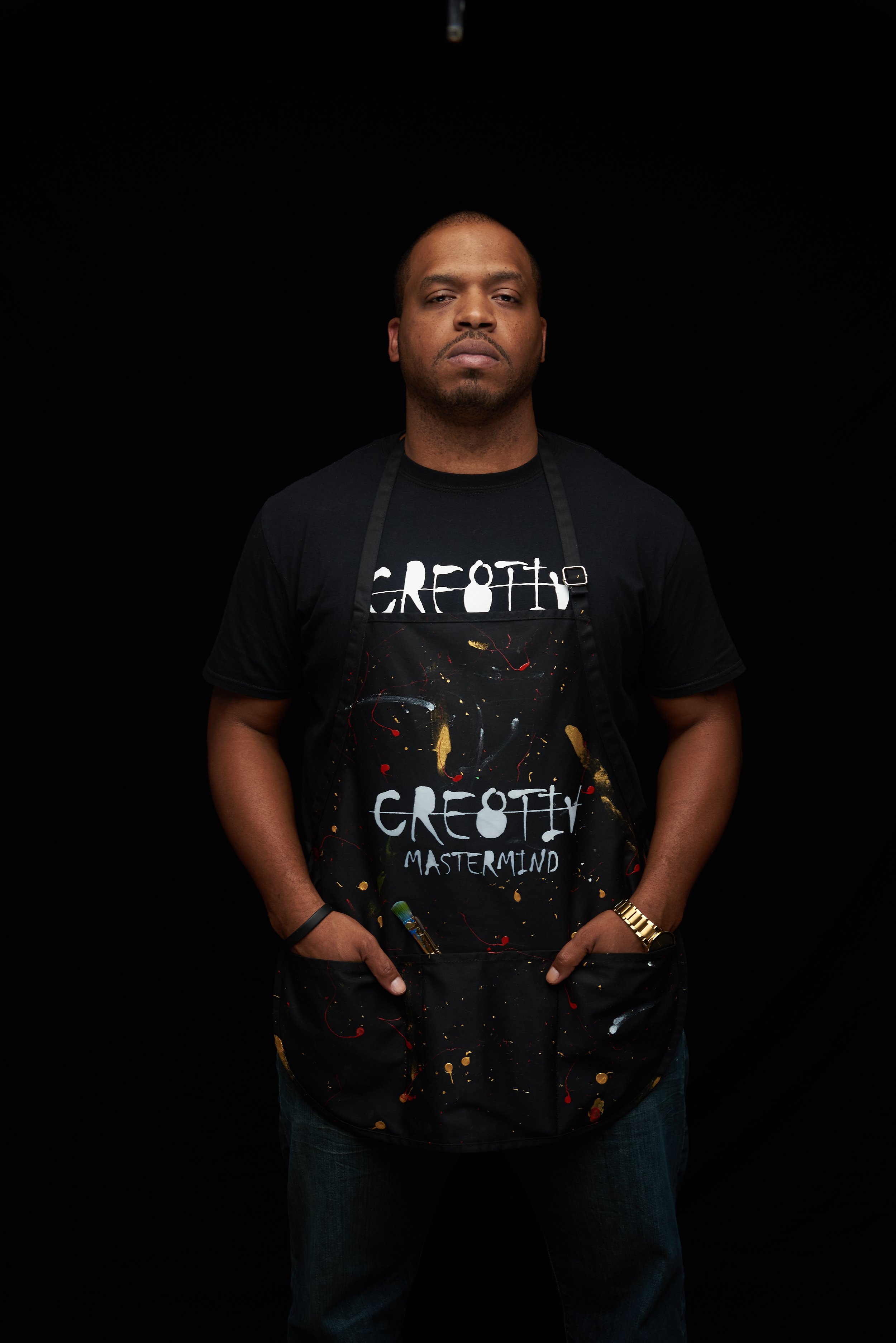 A man wearing a black T-shirt with the words 'GREATH MASTERMIND' and an apron covered in colorful paint splatters, standing against a plain black background.