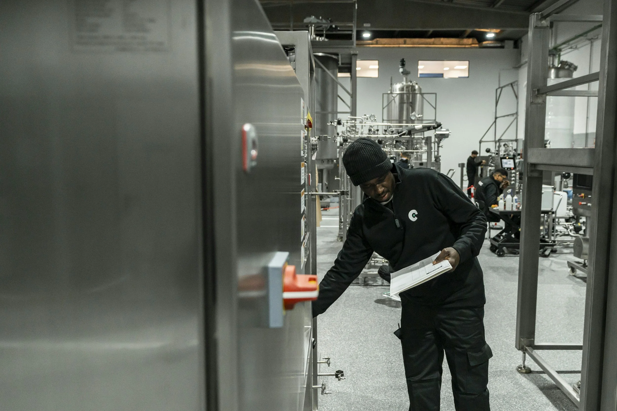 A person wearing a black beanie and black jacket reading a booklet in a room filled with industrial machinery and equipment, with other people working in the background.