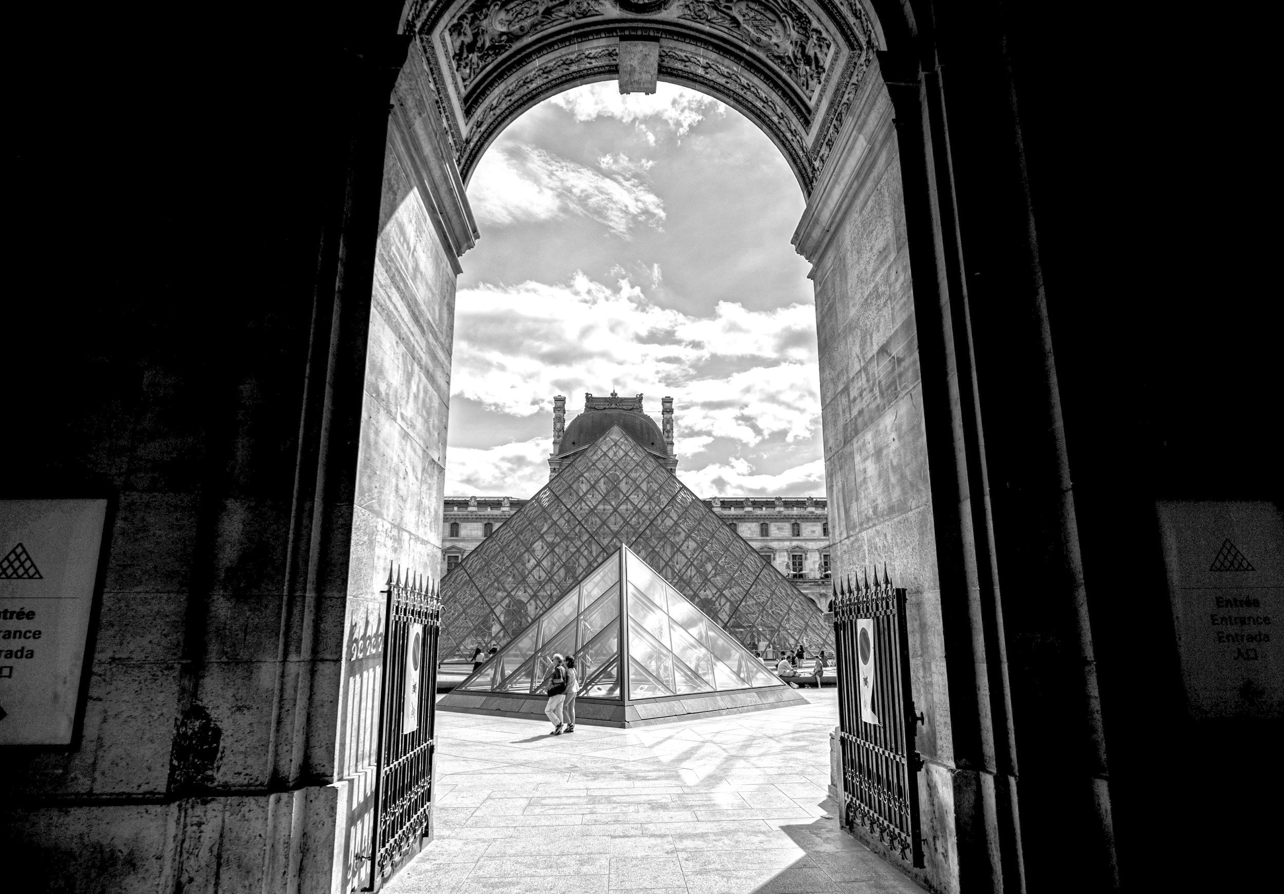 View of the entrance to the Louvre Museum in Paris, framed by an ornate archway, with the glass pyramid and historic museum buildings visible in the background, taken on a sunny day.