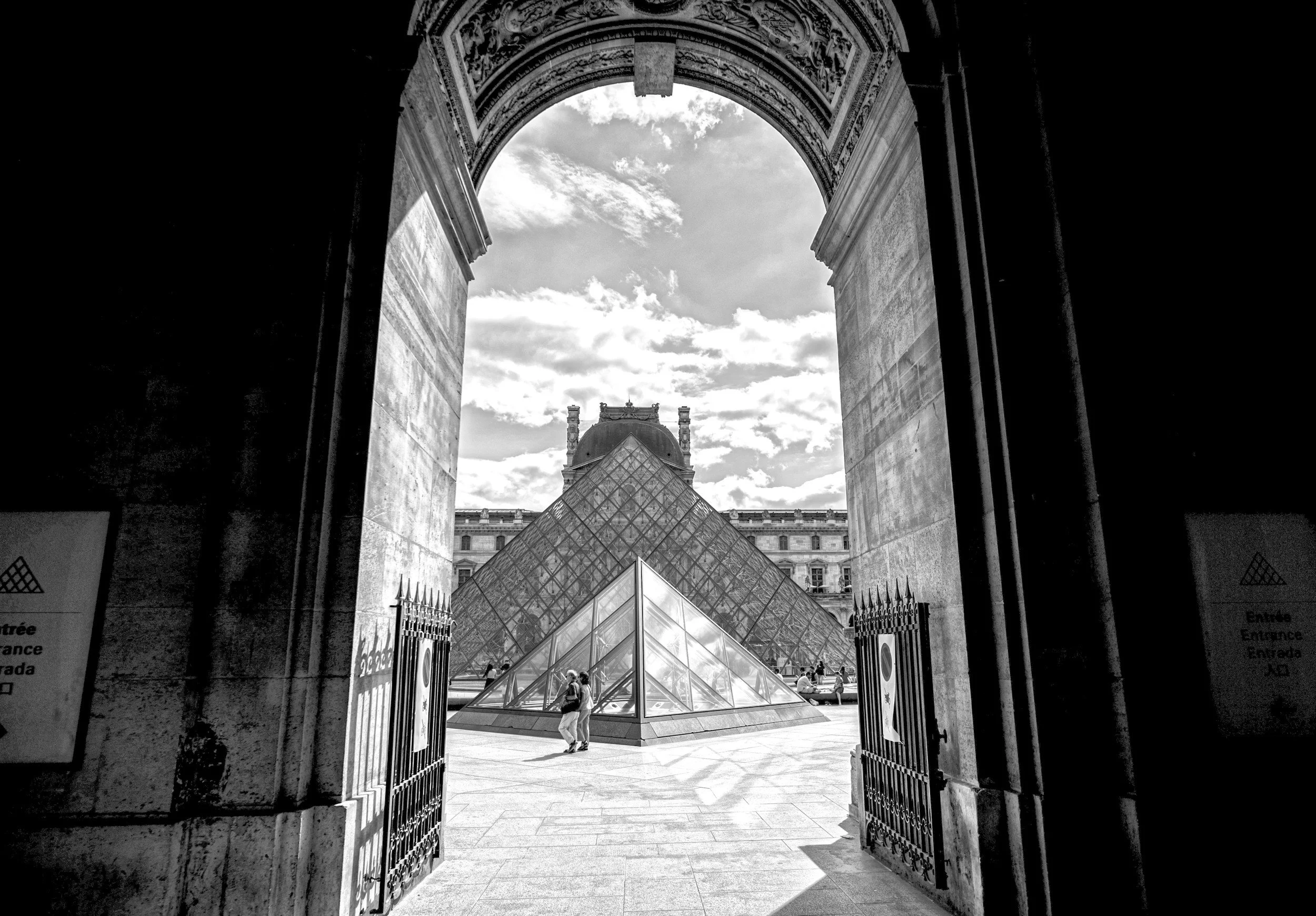 Black and white photo of the glass pyramid at the entrance of the Louvre Museum in Paris, viewed through an arched stone doorway.