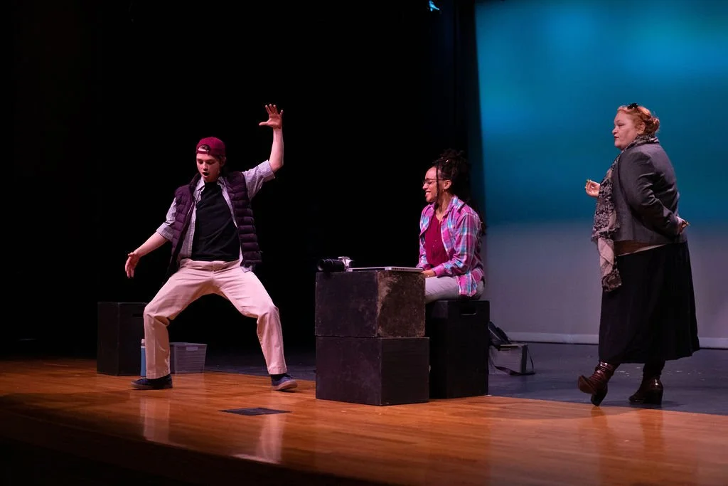 Three actors performing a play called Gracie Vs The People on stage, one standing with her hands in her pockets, another sitting at a desk and smiling, the third standing with arms waving.