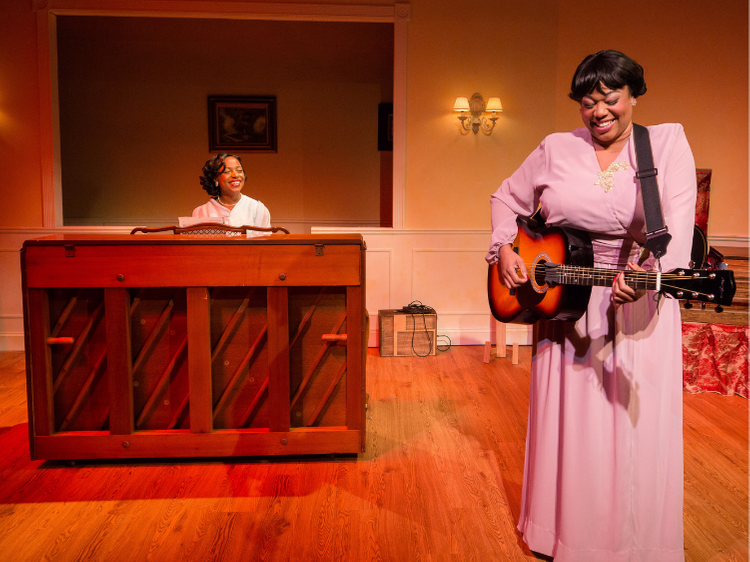 Two women performing in a vintage-style room; one playing piano, the other singing and playing guitar, both smiling.