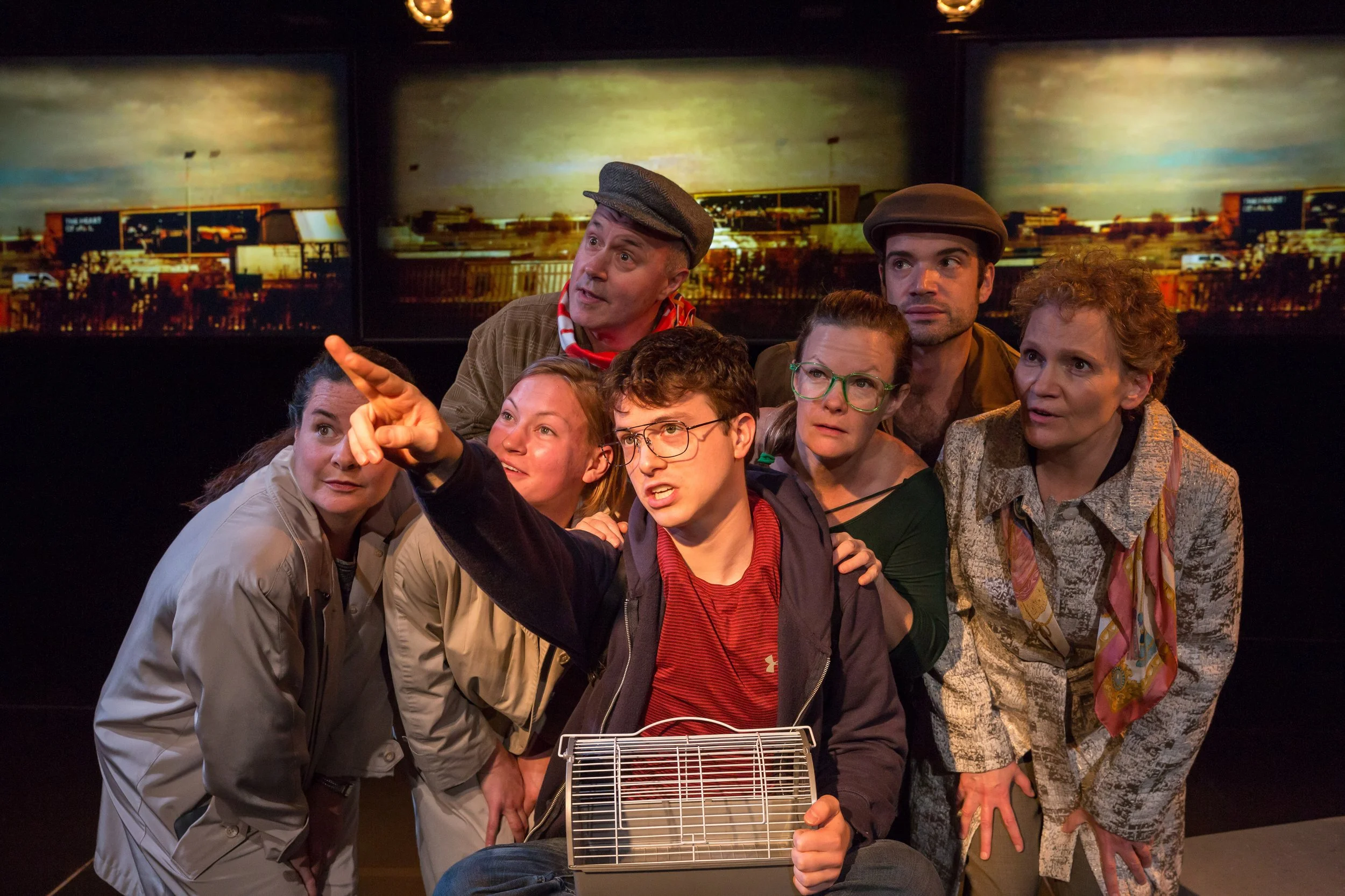 A group of people gathered closely together in a room, looking and pointing at something off-camera. They seem engaged and intrigued, with a background of two large, abstract cityscape murals.
