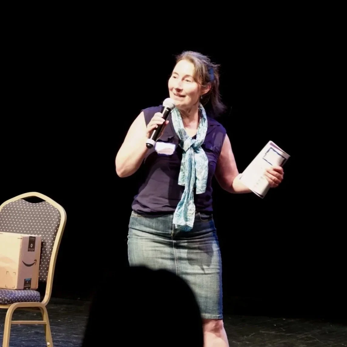 A woman standing on a stage holding a microphone in one hand and a booklet in the other, with a black background and a chair with a cardboard box on it nearby.