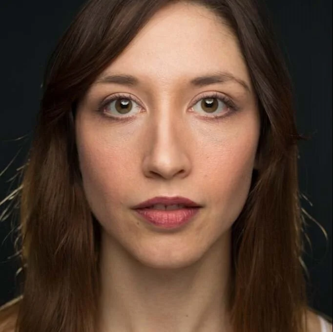 Close-up portrait of a woman with light skin, long brown hair, and blue eyes, looking directly at the camera with a neutral expression, dark background.