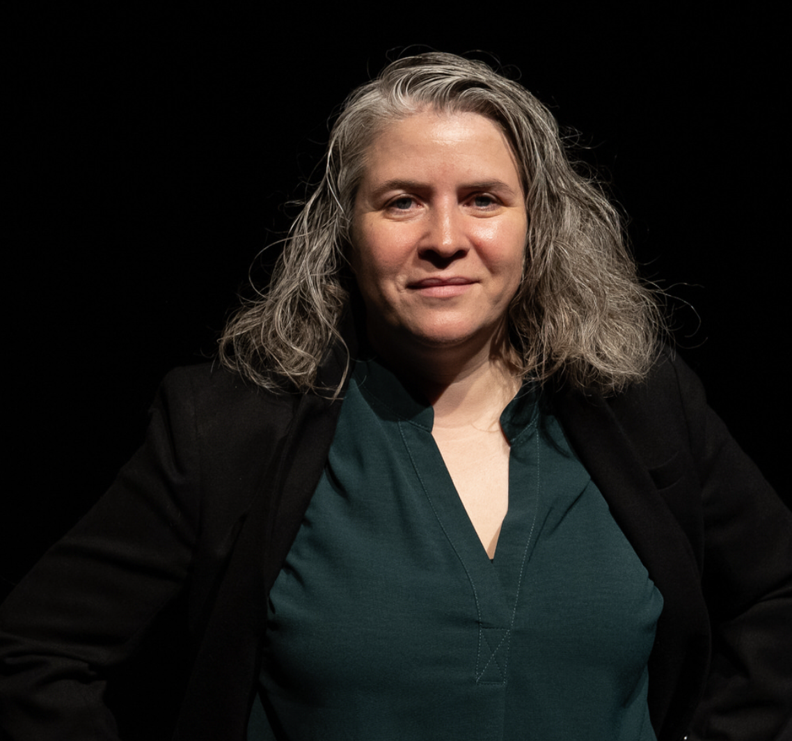 A woman with shoulder-length gray hair wearing a dark blazer over a green V-neck top standing against a black background.